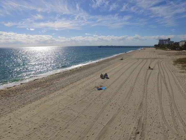 Una amplia playa de arena con huellas y surcos de neumáticos, un muelle a lo lejos, y algunas personas y sombrillas.