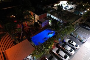 Zona de piscina en la azotea con piscina azul, rodeada de palmeras y algunos coches estacionados.