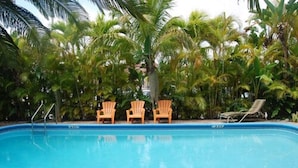Una piscina con tres sillas de madera y una tumbona, rodeada de palmeras.