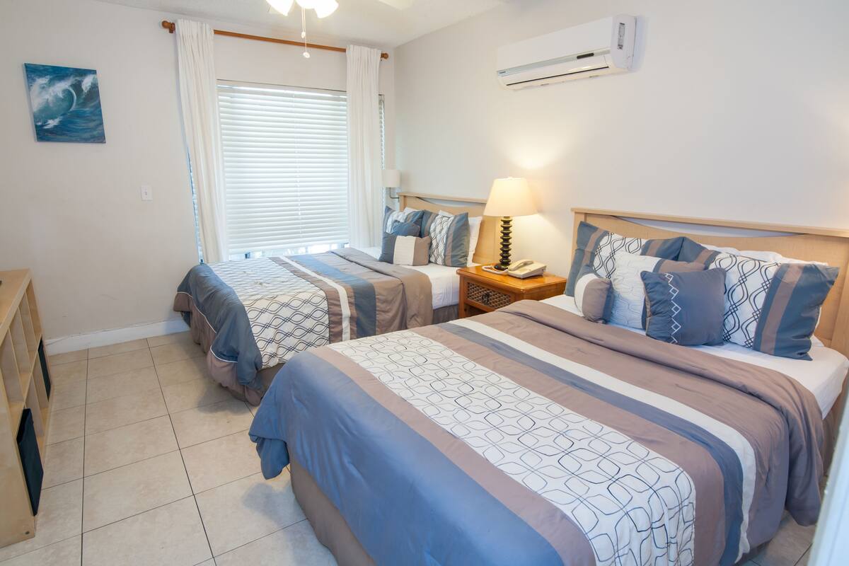 A hotel room with two beds, a bedside table with a lamp, an air conditioning unit, and a window with curtains.