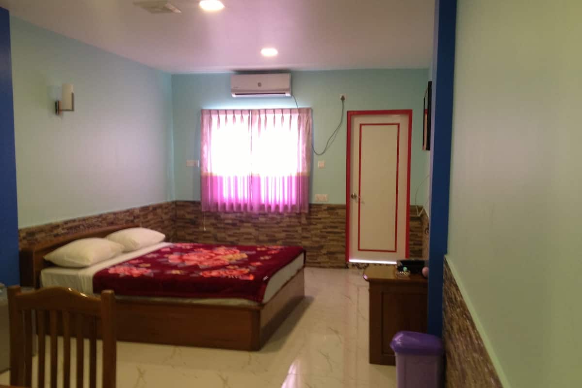 A bedroom with a bed, a wooden chair, a nightstand, a trash bin, a door, and a window with curtains.