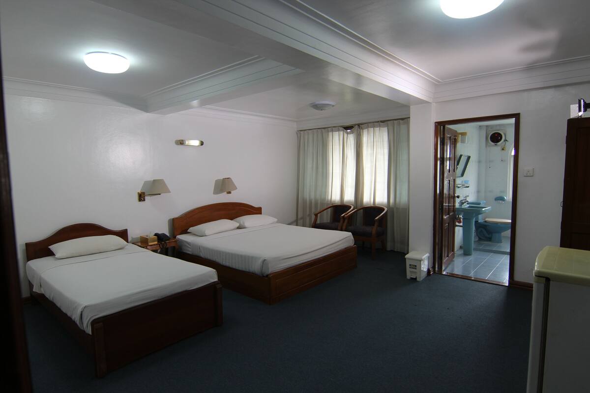 A hotel room with two single beds, a wooden headboard, a small table with a lamp, a chair, and a view of the bathroom through a door.