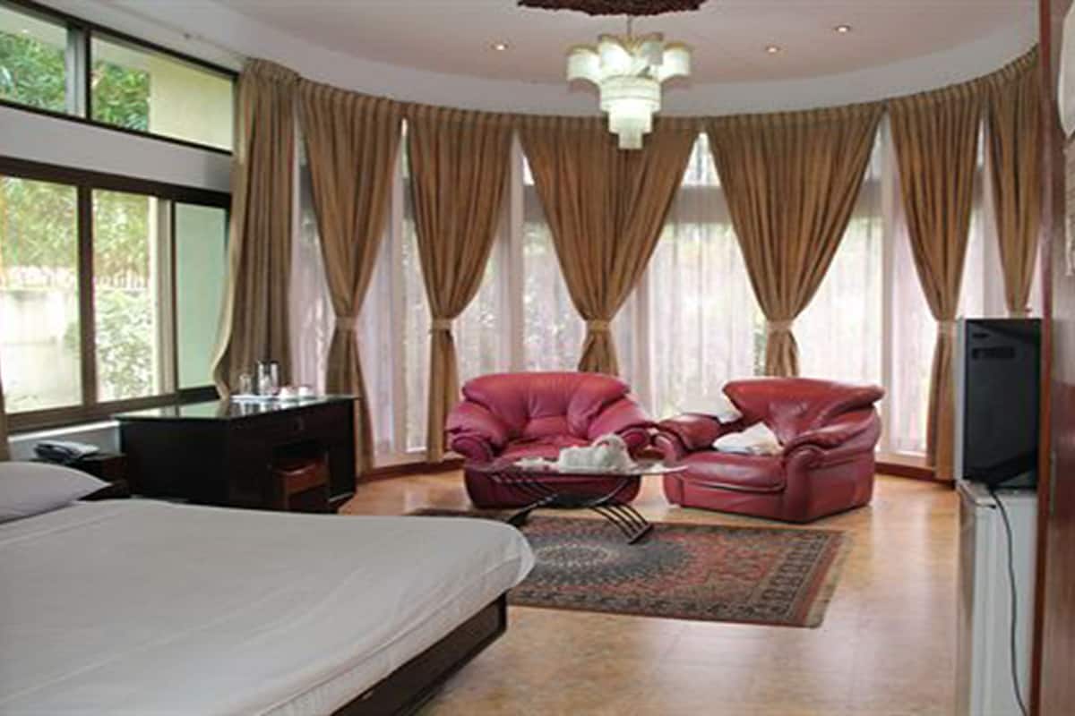 A hotel room with a bed, a desk, a red leather chair, a television, and large windows with curtains.