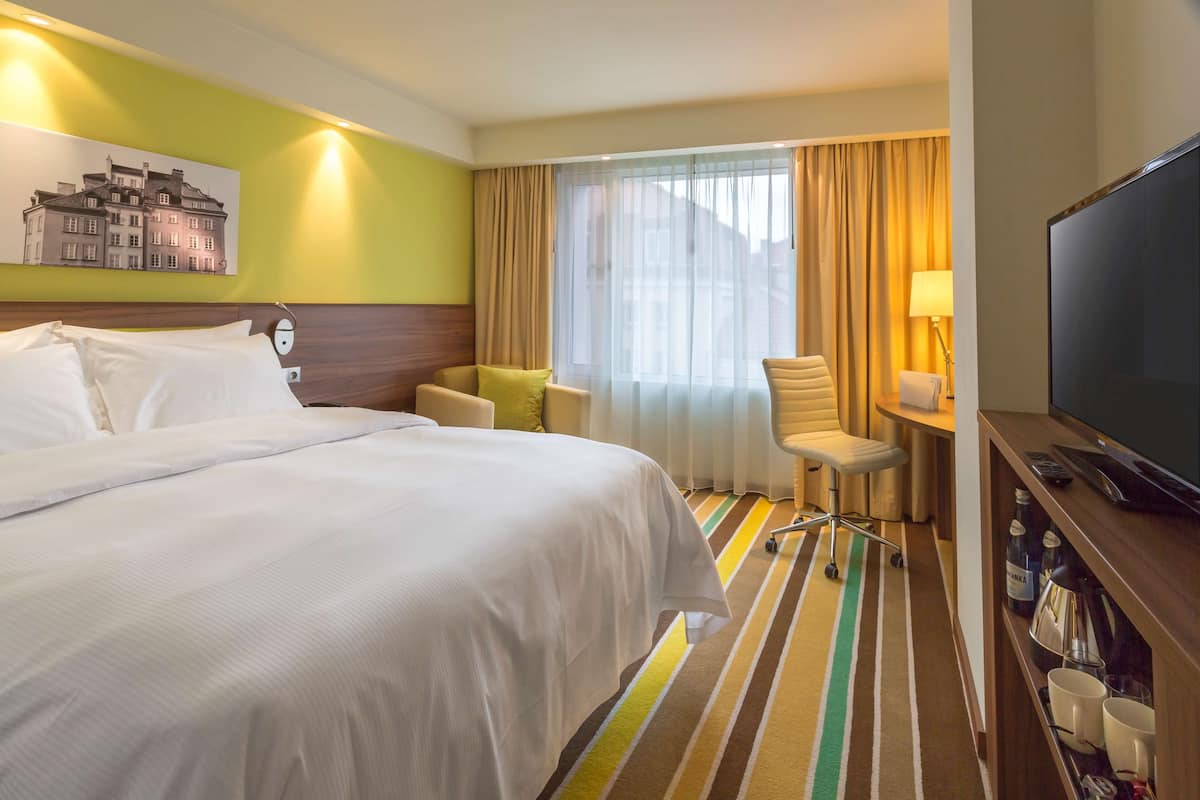 Une chambre d’hôtel avec un grand lit, un bureau avec une chaise, une télévision et une fenêtre avec des rideaux.