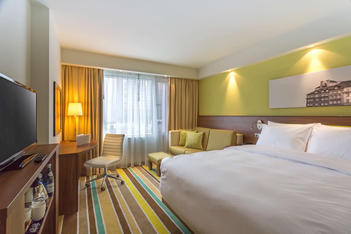 Une chambre d’hôtel avec un grand lit, un bureau avec une télévision, une chaise, un canapé et une fenêtre avec des rideaux.