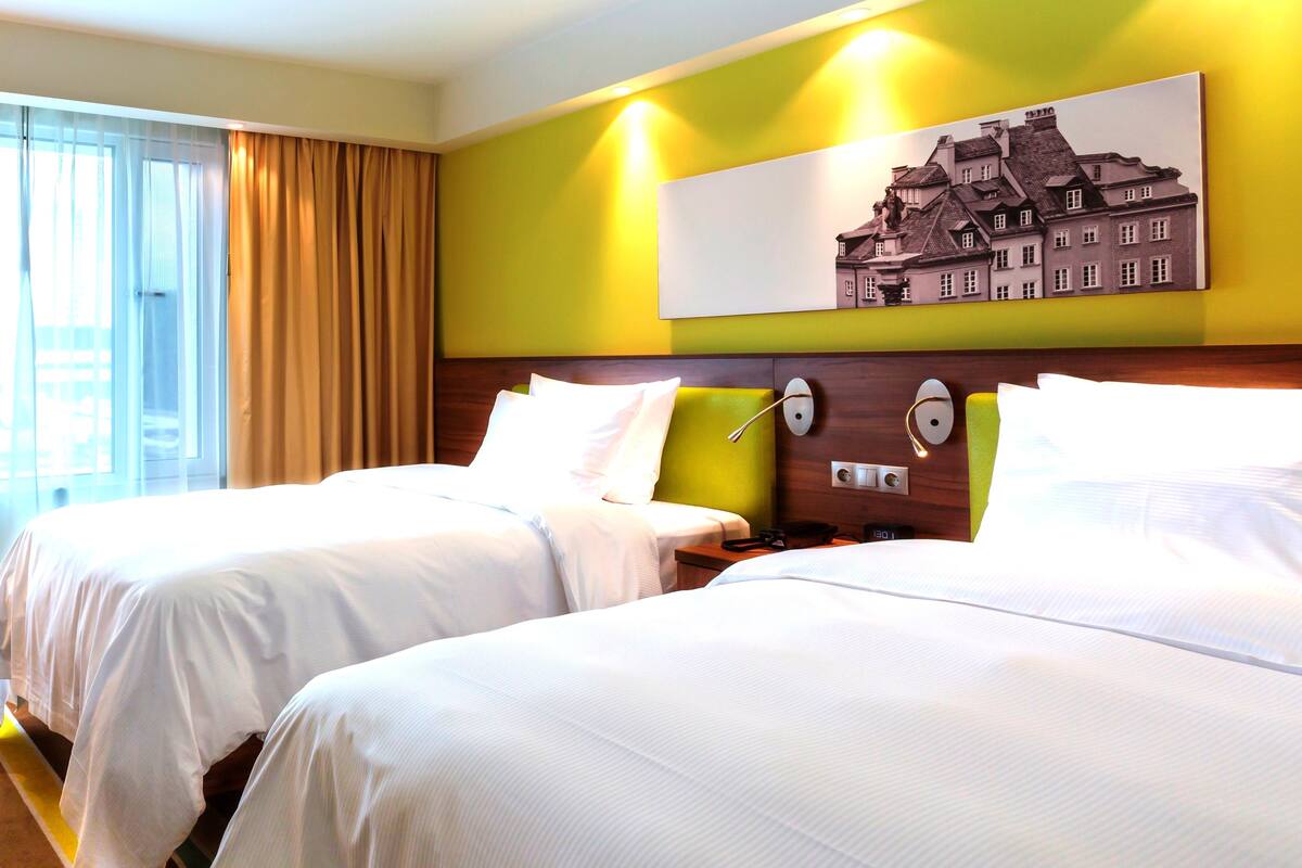 Une chambre d’hôtel avec deux lits, une grande fenêtre avec des rideaux, un tableau accroché au mur et une table de chevet avec une lampe.