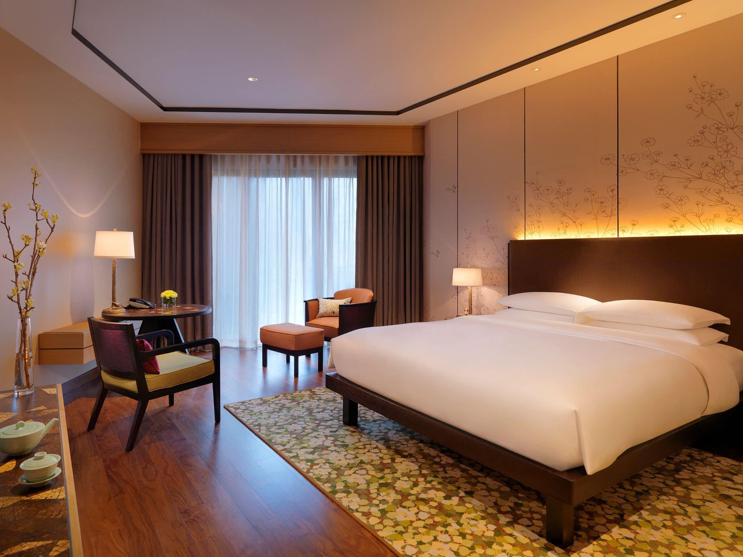 A hotel room with a large bed, a desk with a chair, a small table with a lamp, and a vase with flowers.