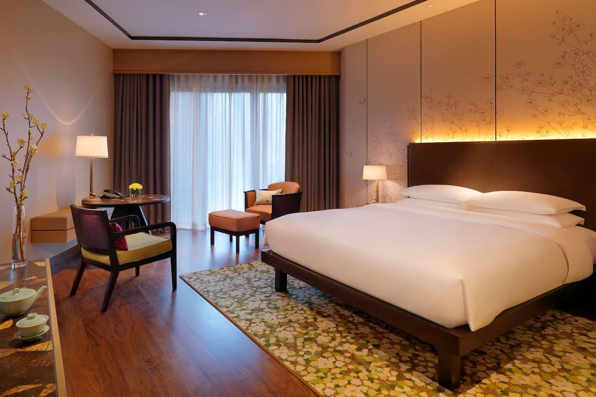 A hotel room with a large bed, a desk with a chair, a small table with a lamp, and a vase with flowers.