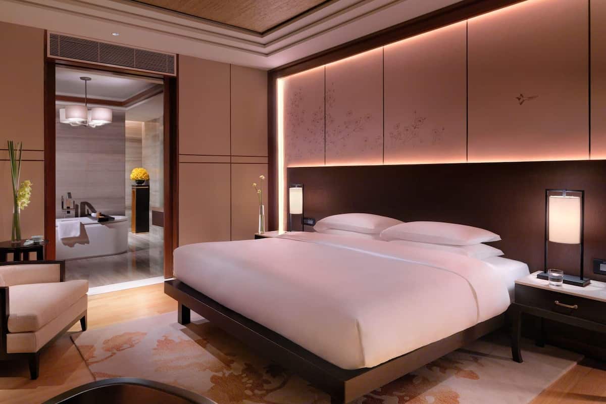 A modern hotel room with a large bed, bedside lamps, a chair, and a bathroom visible through an open door.