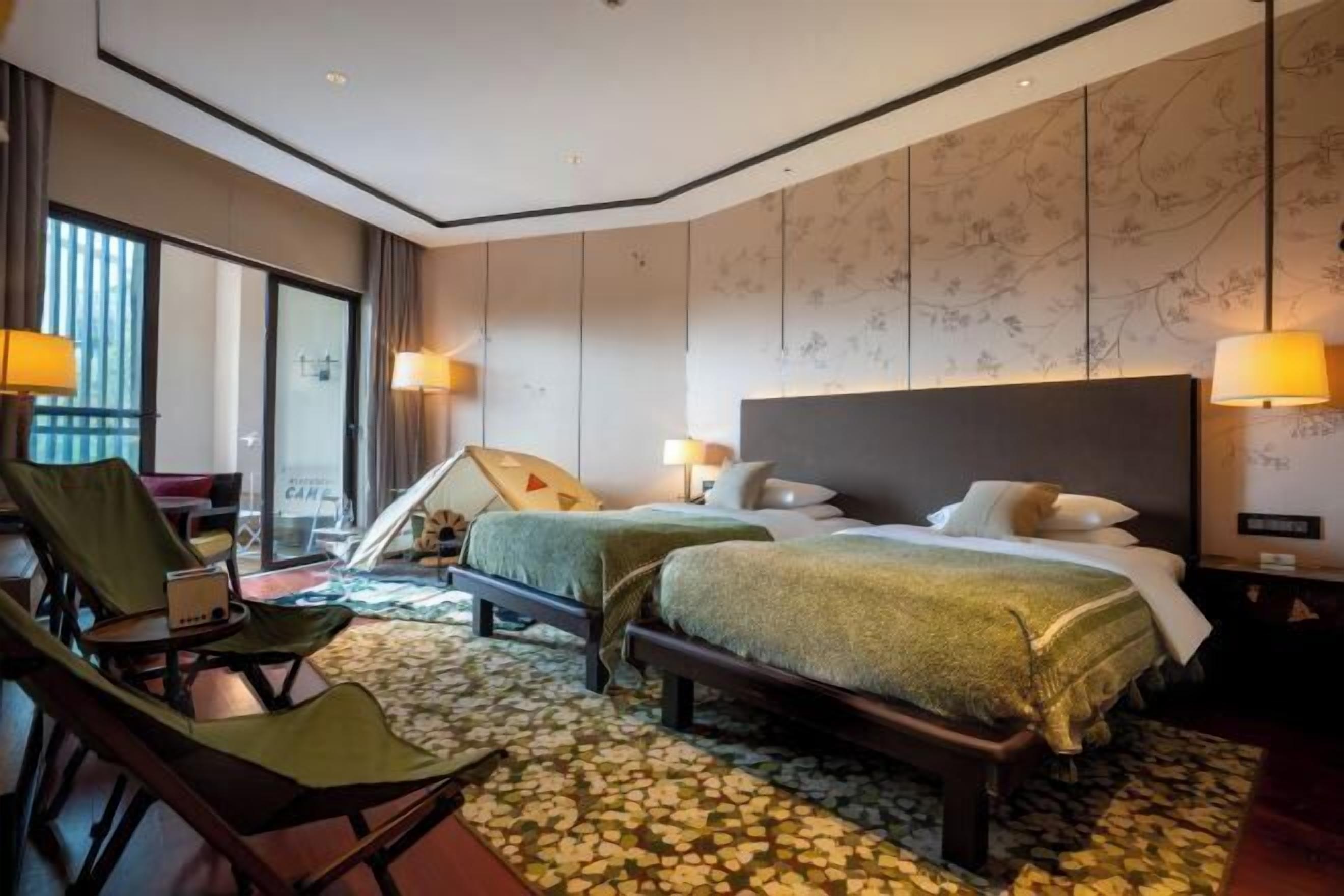 A hotel room with two beds, a sofa, and a chair. There are lamps on either side of the beds and a large window with curtains.