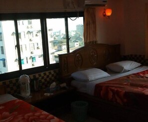 A hotel room with two beds, a window with a view of buildings, a nightstand with a lamp, and a wall-mounted air conditioner. - M.G.M Hotel (Yangon)