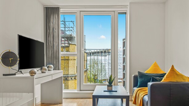 Een moderne woonkamer met een groot raam, een flatscreen televisie, een kleine tafel en een bank met kussens.