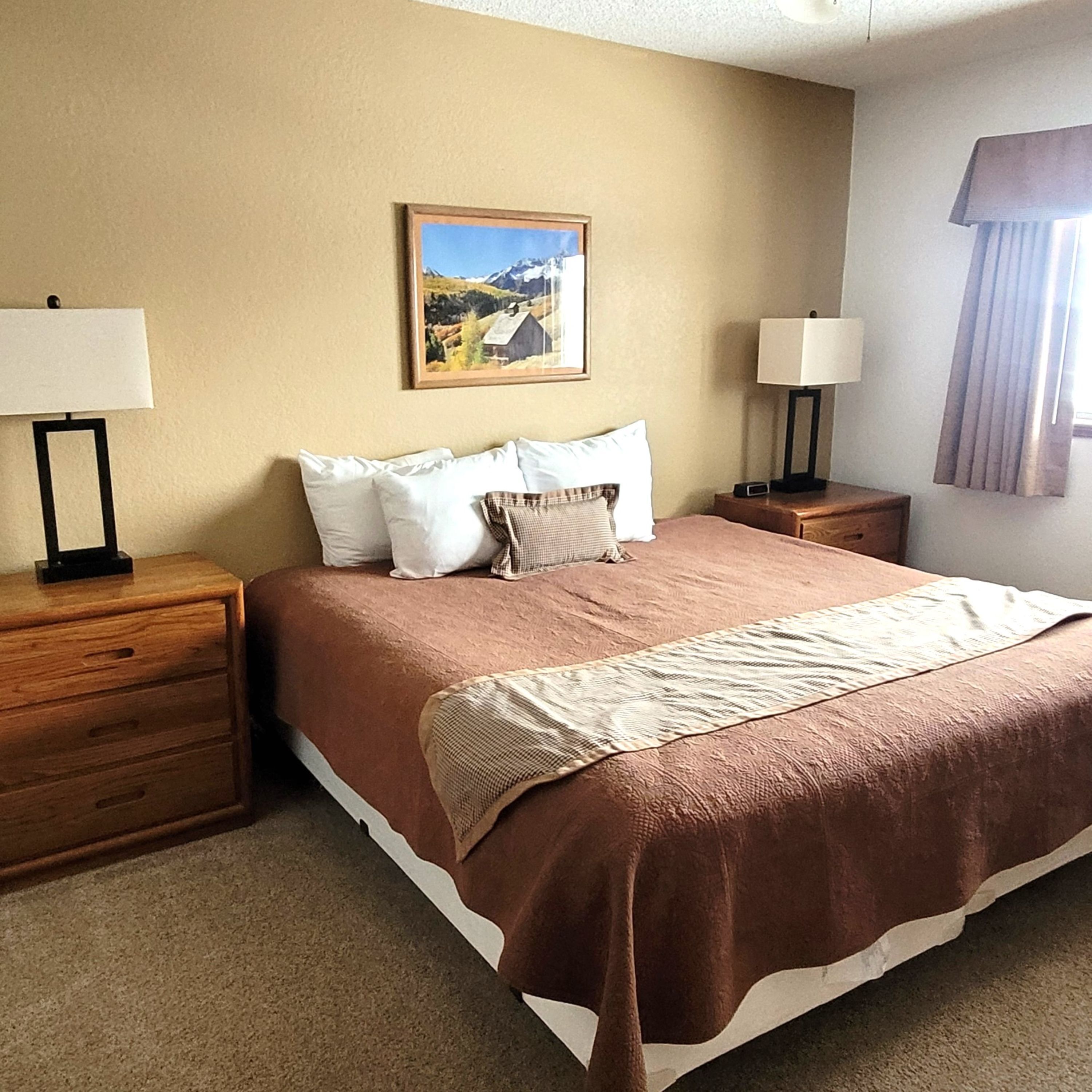Une chambre d’hôtel avec un grand lit, deux tables de chevet, une commode, une lampe, un tableau au mur et une fenêtre avec des rideaux.