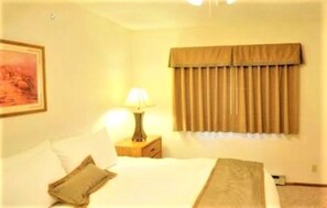 A hotel room with a bed, a nightstand with a lamp, a window with curtains, and a painting on the wall.