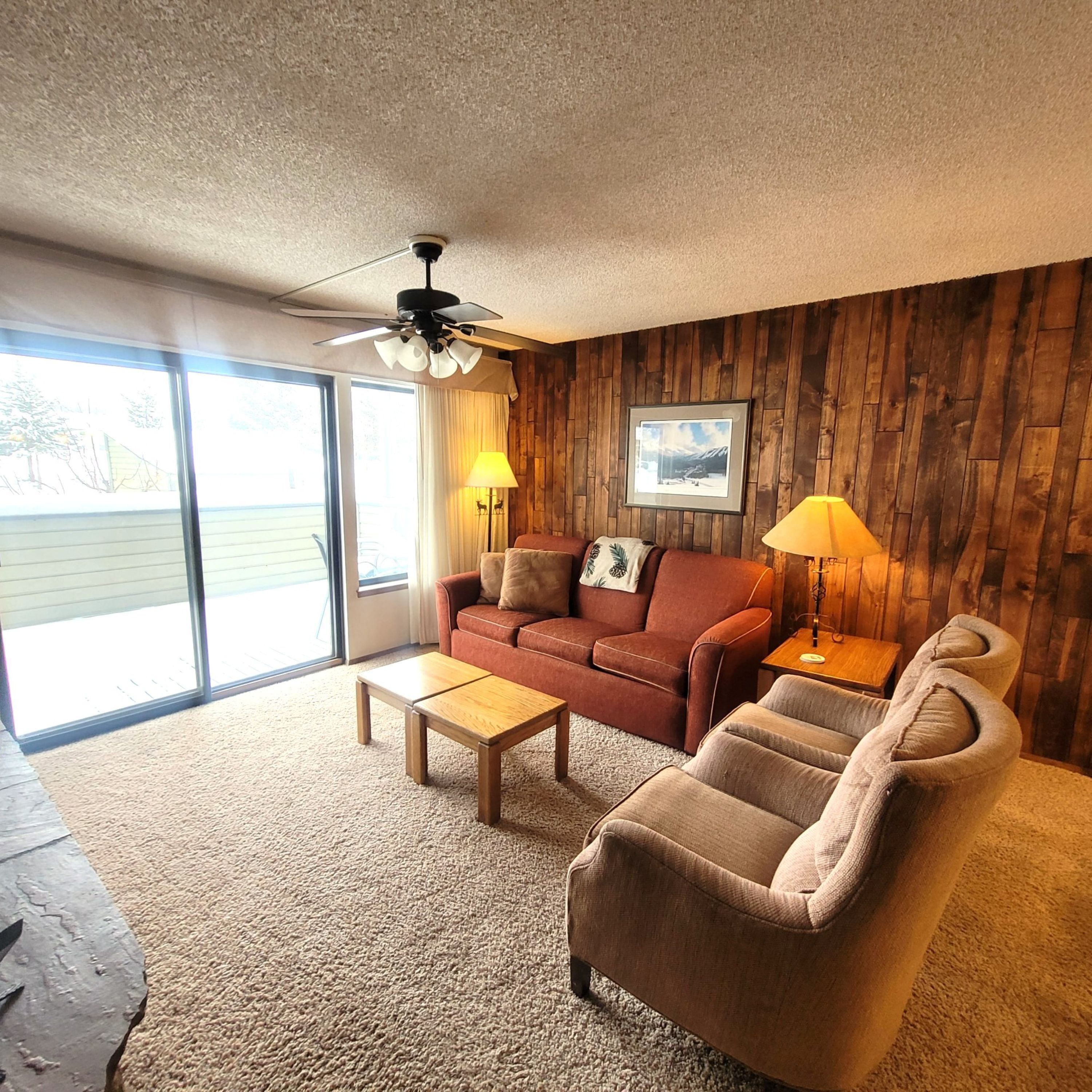 A cozy living room with a wooden wall, a ceiling fan, a sofa, a chair, a coffee table, and a lamp.