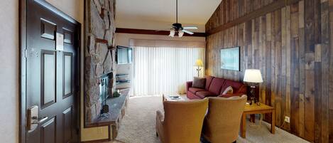 A hotel room with a stone wall, a wooden panel wall, a ceiling fan, a TV, a sofa, two armchairs, a coffee table, and a lamp.
