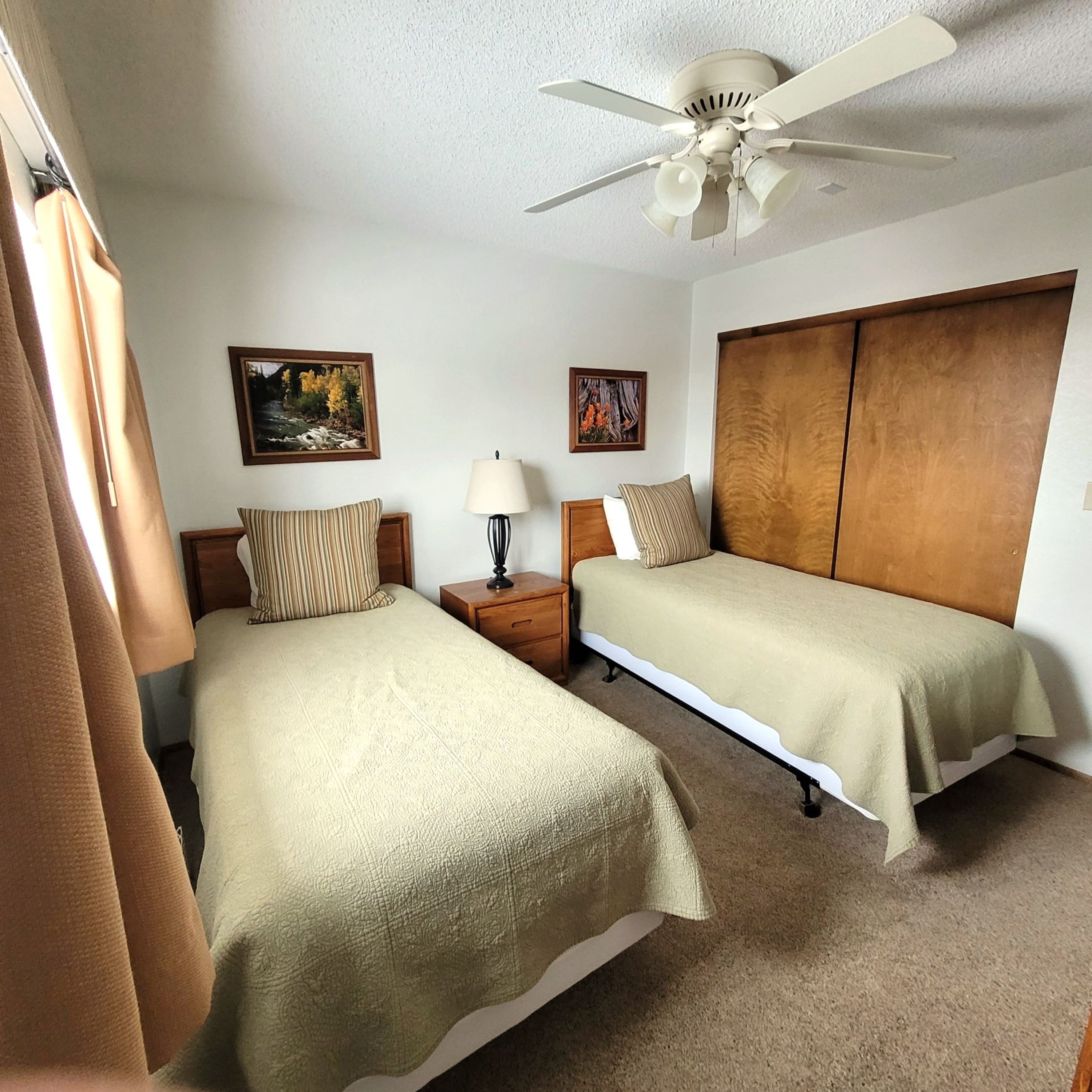 A bedroom with two single beds, a ceiling fan, a nightstand with a lamp, and two framed pictures on the wall.