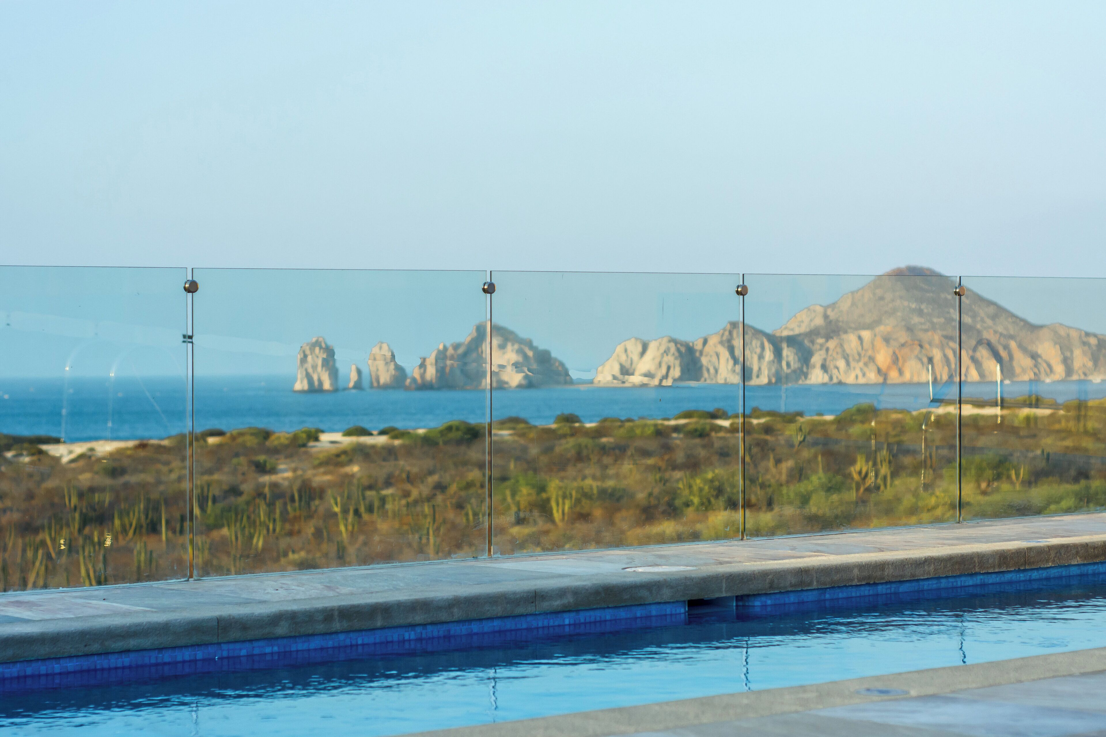 A swimming pool with clear glass panels, a view of the sea, and rocky formations in the distance.