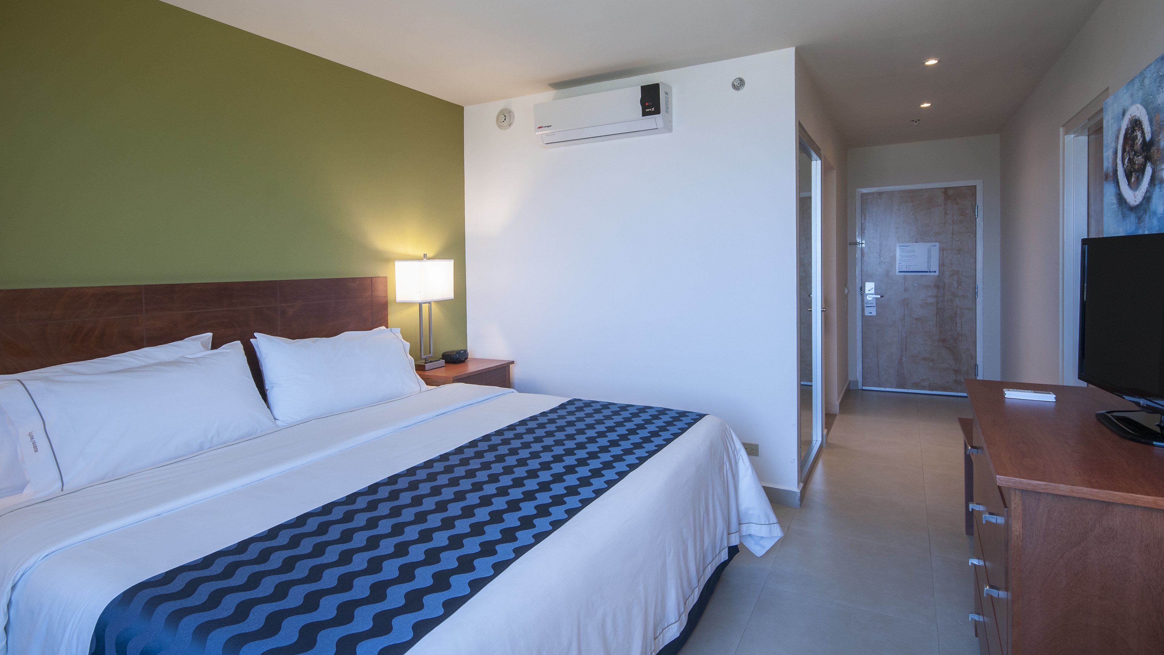 A hotel room with a bed, a bedside lamp, a TV, and a door leading to another room.