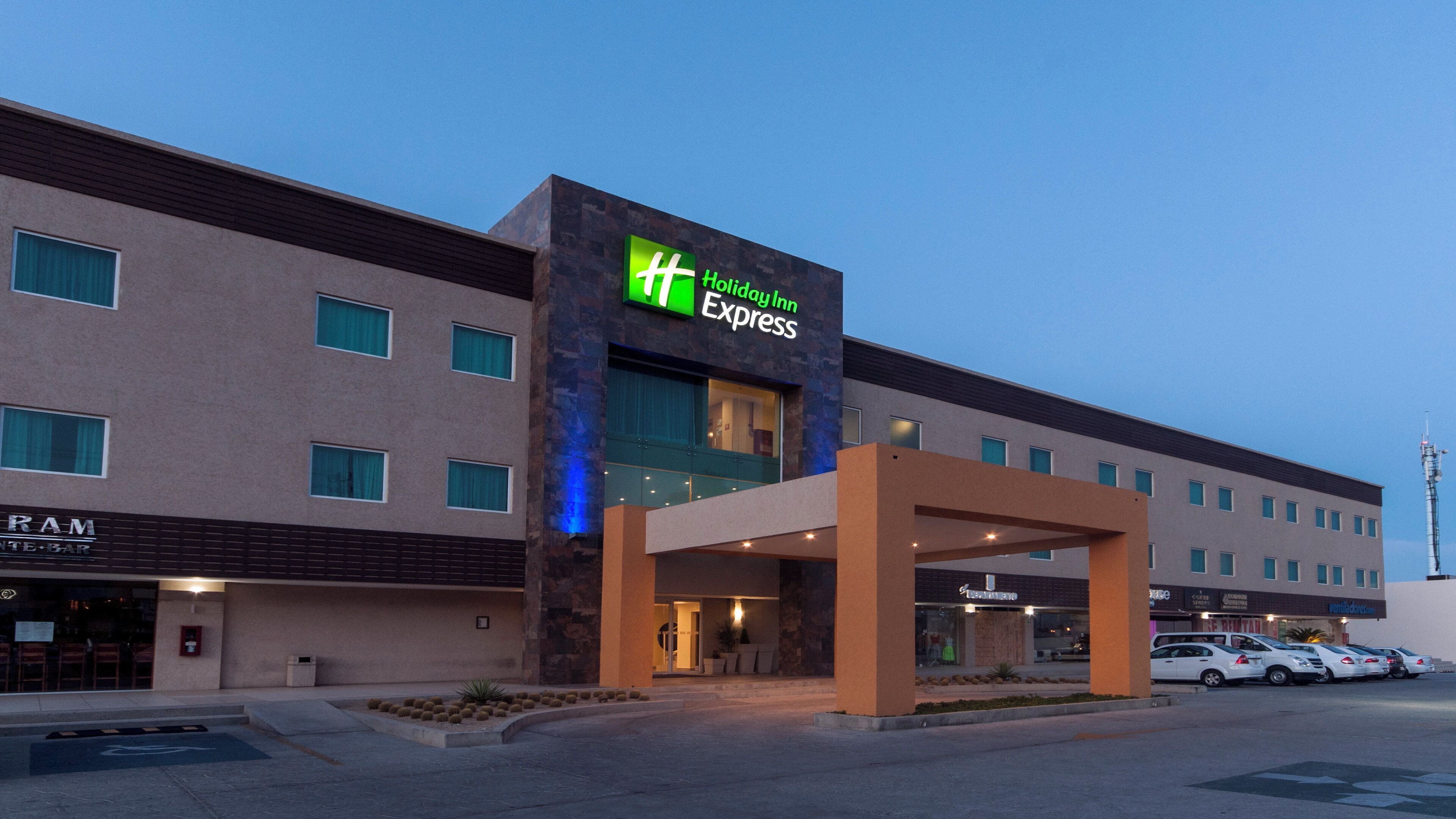 Edificio de Holiday Inn Express con diseño moderno, que presenta una entrada destacada y un área de estacionamiento con varios autos.