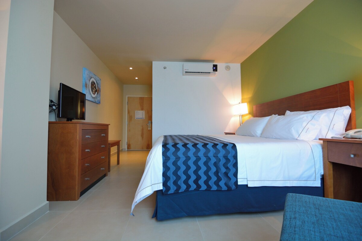 A hotel room with a bed, a desk, a chair, a dresser, a television, and an air conditioning unit.