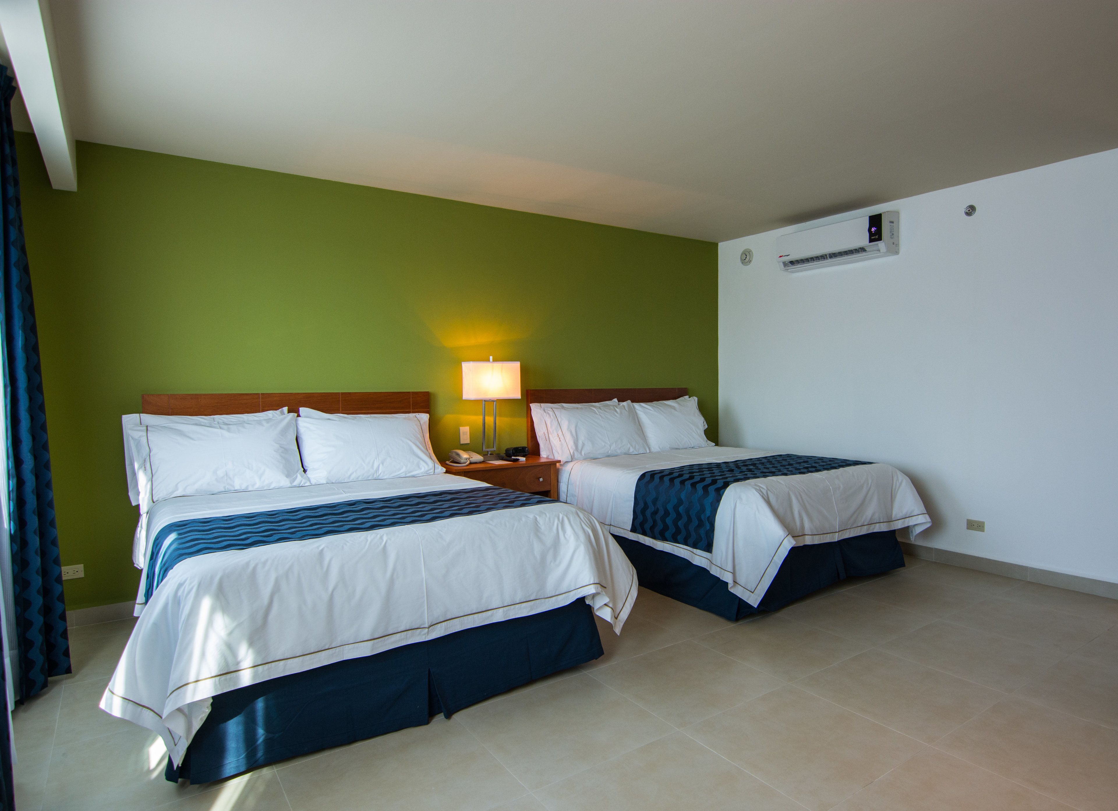 A hotel room with two beds, a nightstand with a lamp, and a wall-mounted air conditioner.