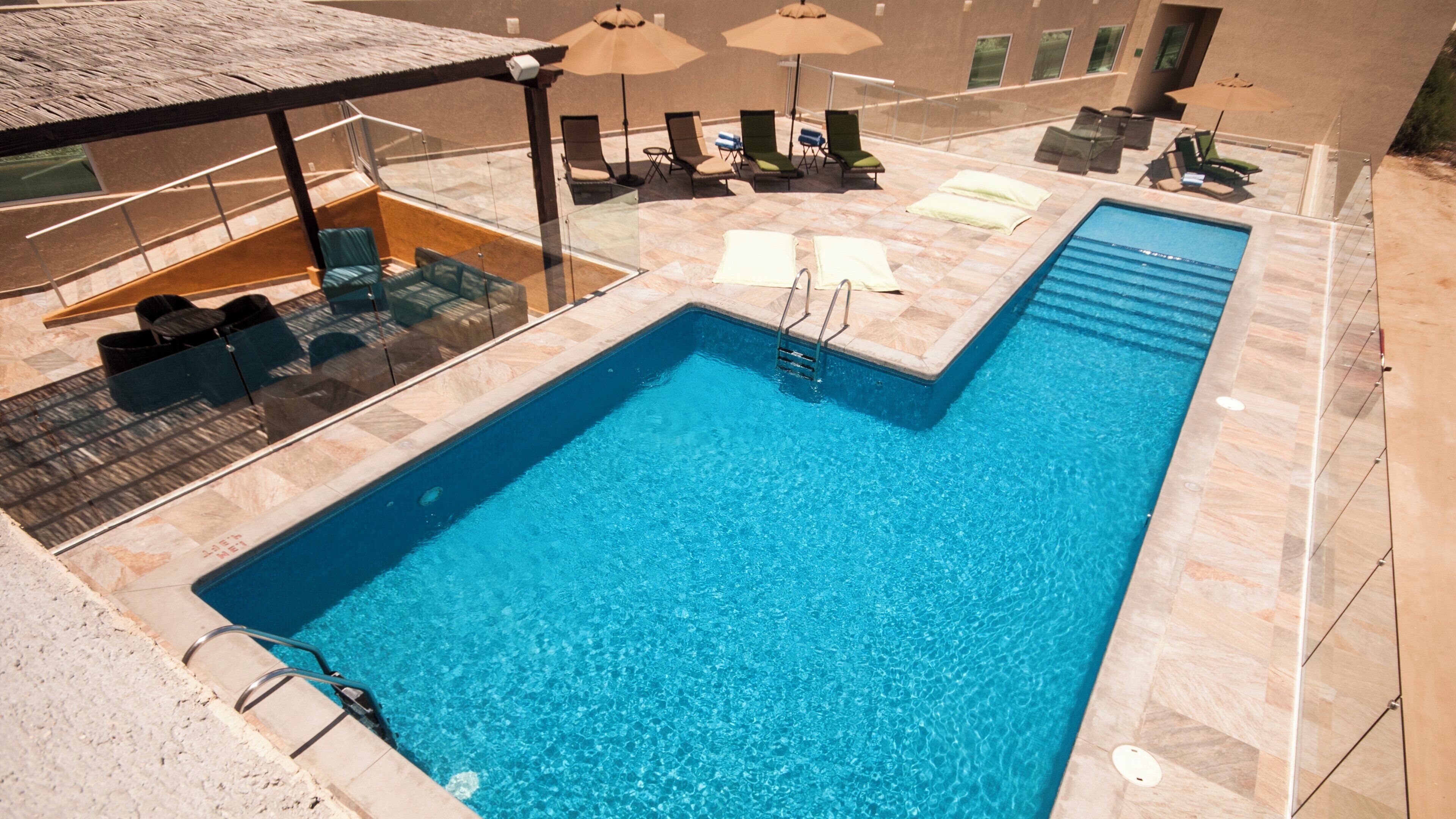A rectangular swimming pool with a clear blue water, surrounded by lounge chairs and umbrellas, and a covered seating area with a glass balustrade.