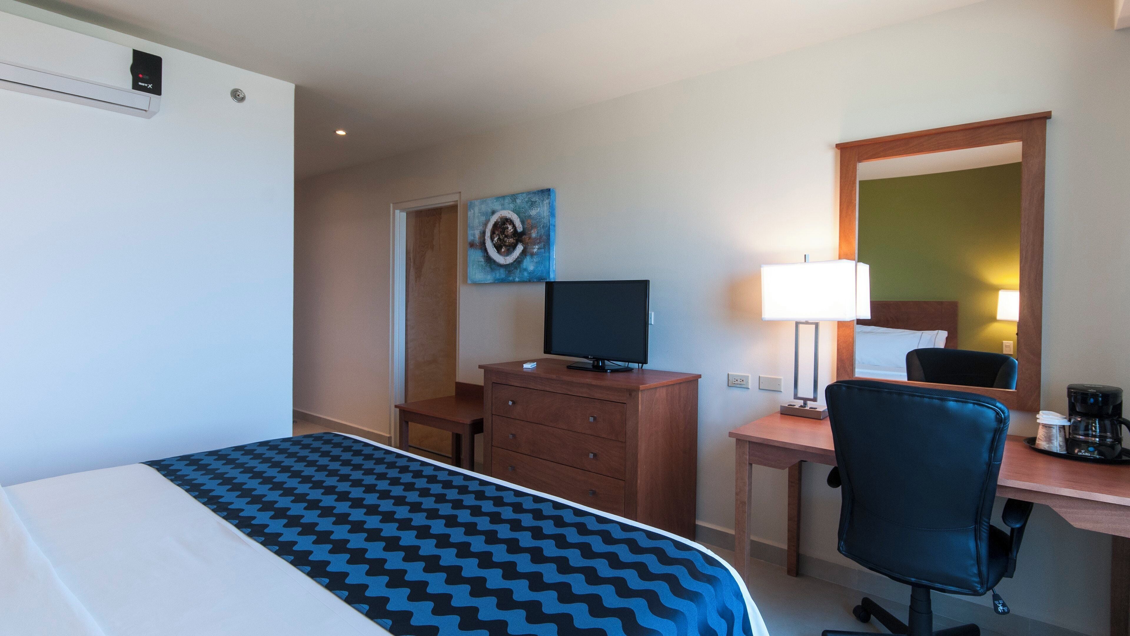A hotel room with a bed, a desk with a computer, a chair, a lamp, and a painting on the wall.