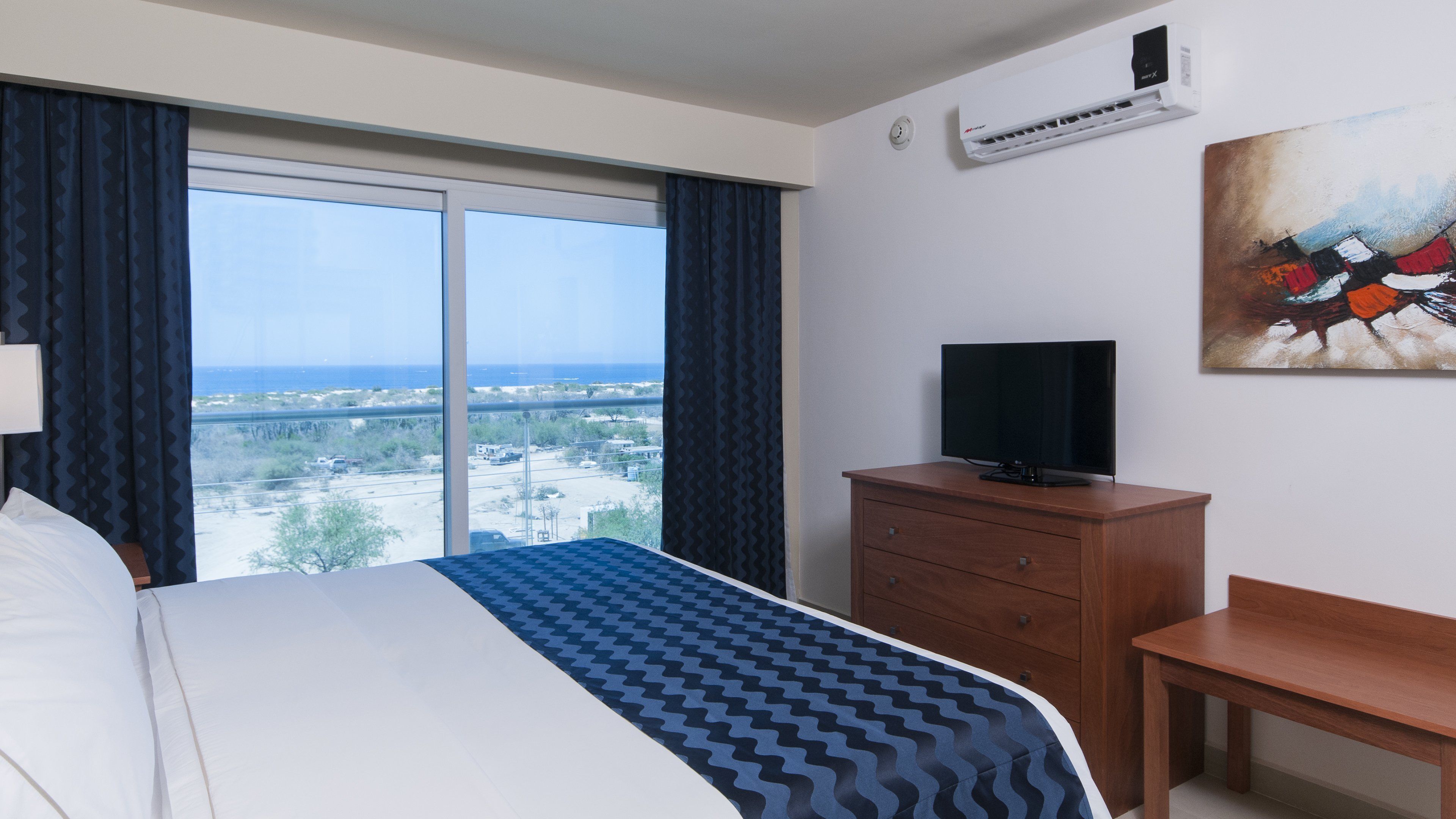 A hotel room with a bed, a TV on a wooden dresser, a painting on the wall, and a view of the city and ocean through the window.