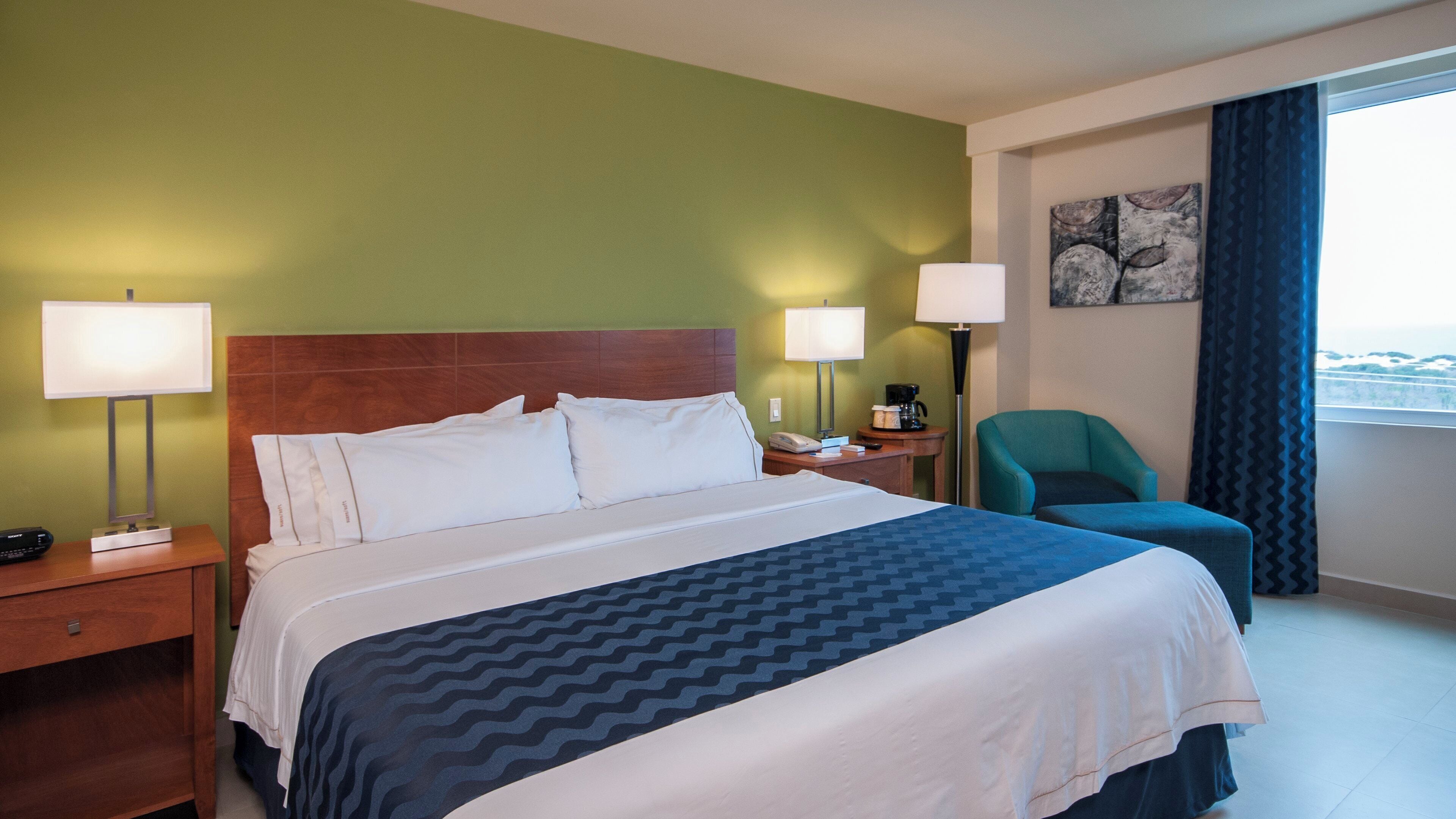 Una habitación de hotel con una cama, dos lámparas de noche, una silla, una mesita de noche, una cafetera y un cuadro en la pared.