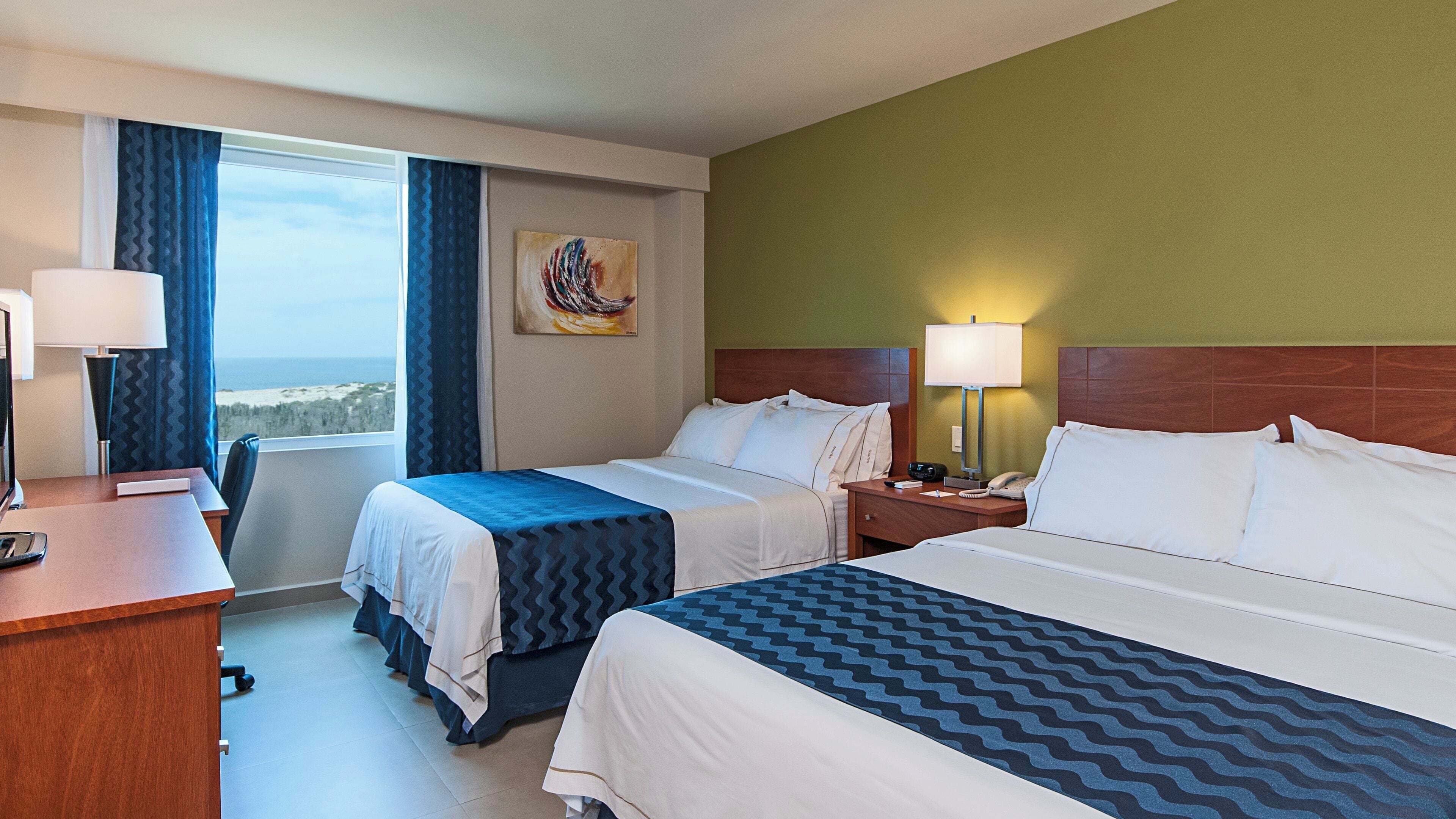A hotel room with two beds, a desk, a chair, a nightstand, and a painting on the wall.
