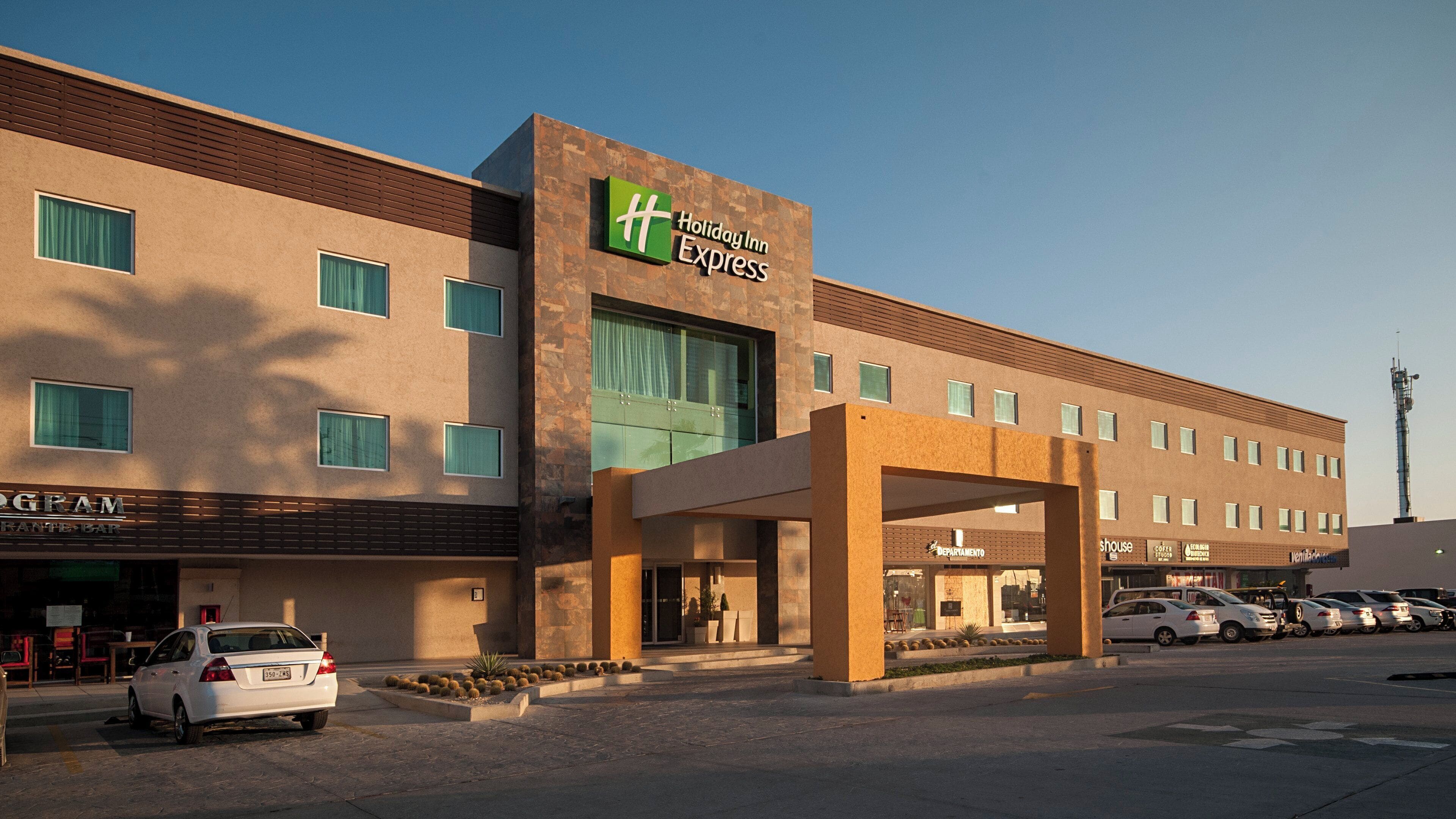 Un bâtiment de l’hôtel Holiday Inn Express, doté d’un design moderne, avec une entrée imposante et un parking où sont stationnées plusieurs voitures.