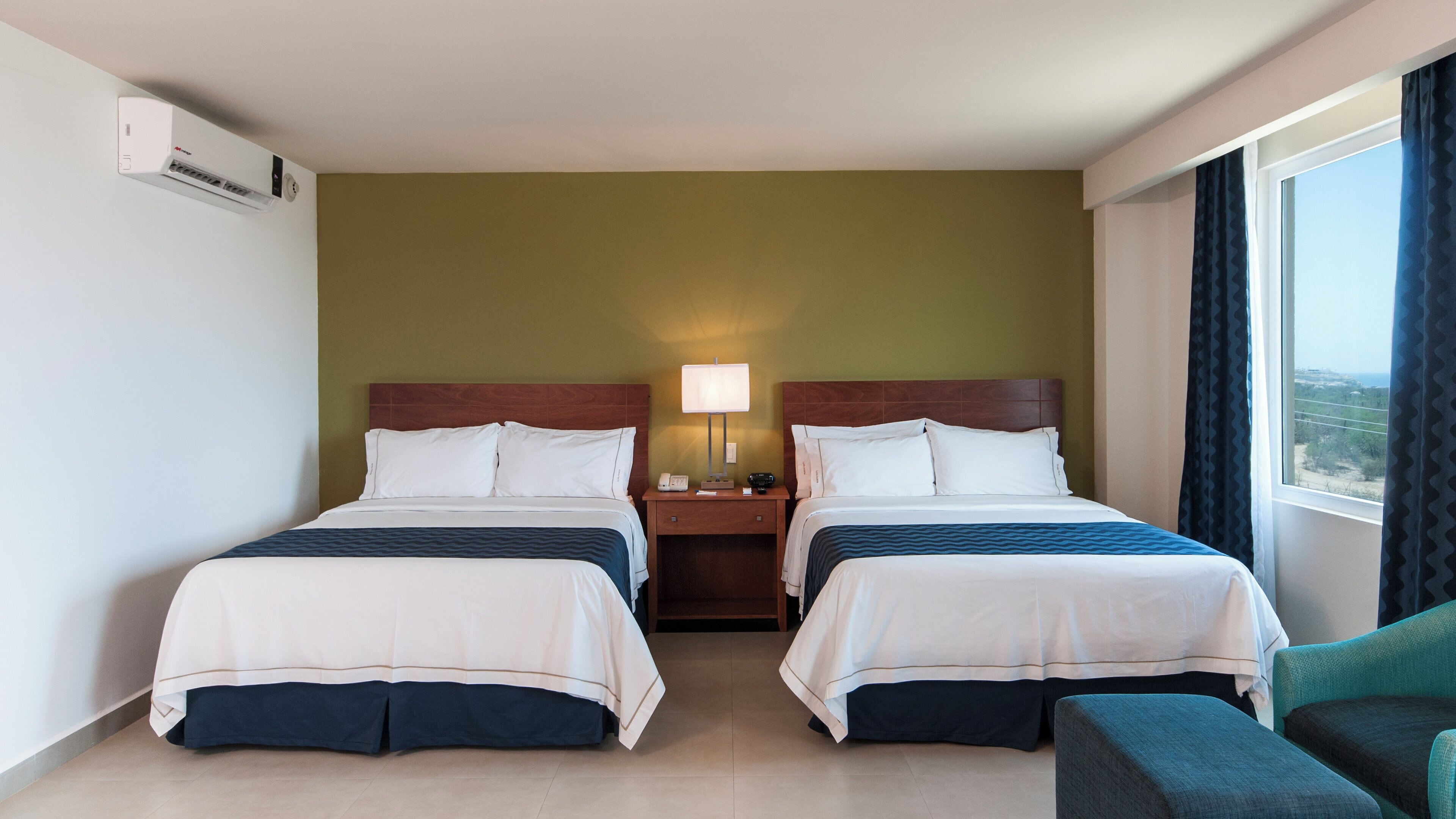 Une chambre d’hôtel avec deux lits, une table de chevet avec une lampe, une chaise et une fenêtre avec des rideaux.
