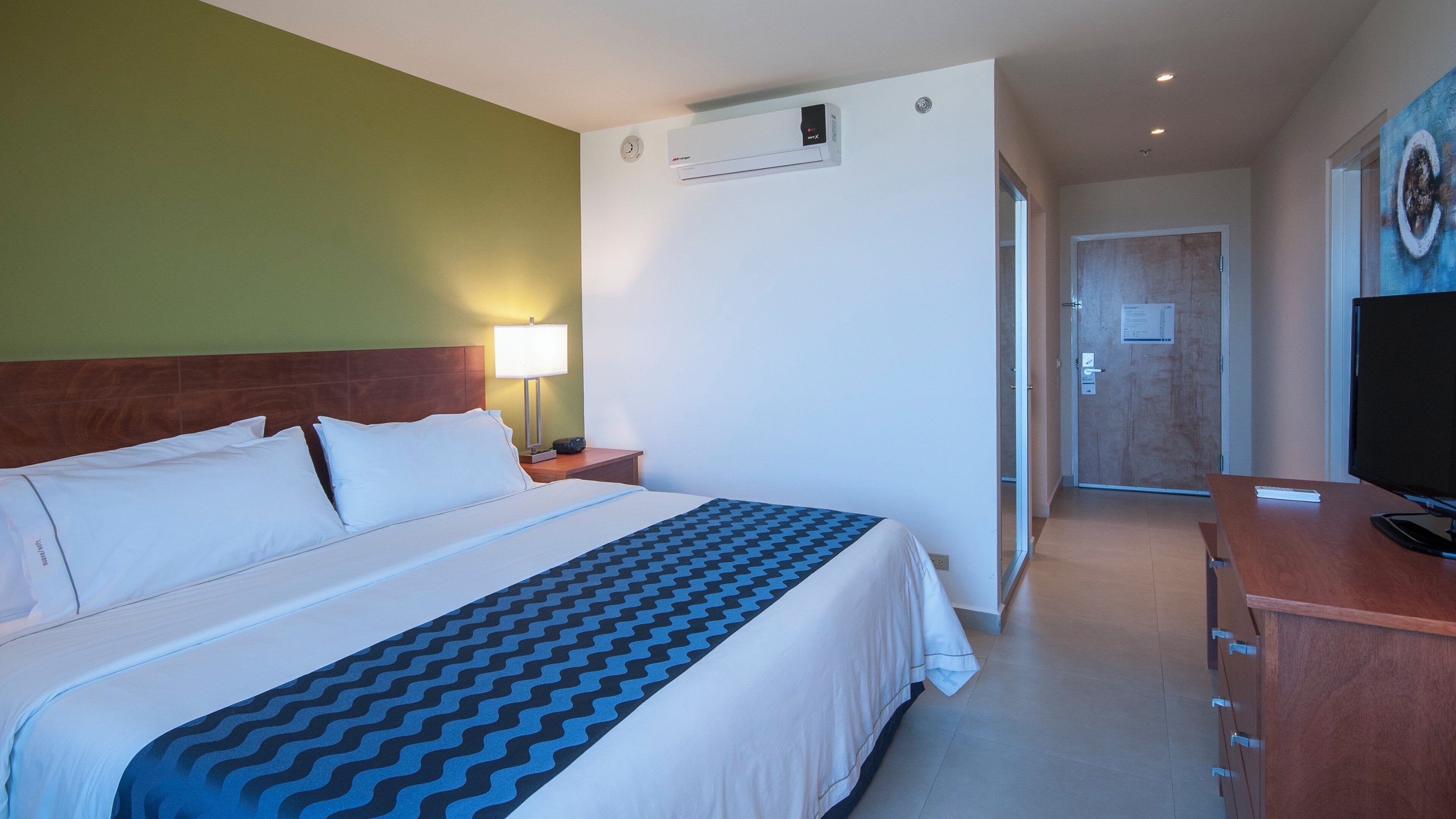 Een hotelkamer met een bed, een bedlamp, een airconditioningsysteem, een televisie en een deur naar een andere kamer.