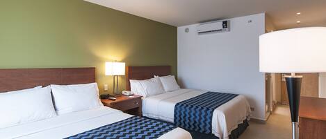 A hotel room with two beds, a nightstand, a lamp, an air conditioning unit, and a desk with a lamp.