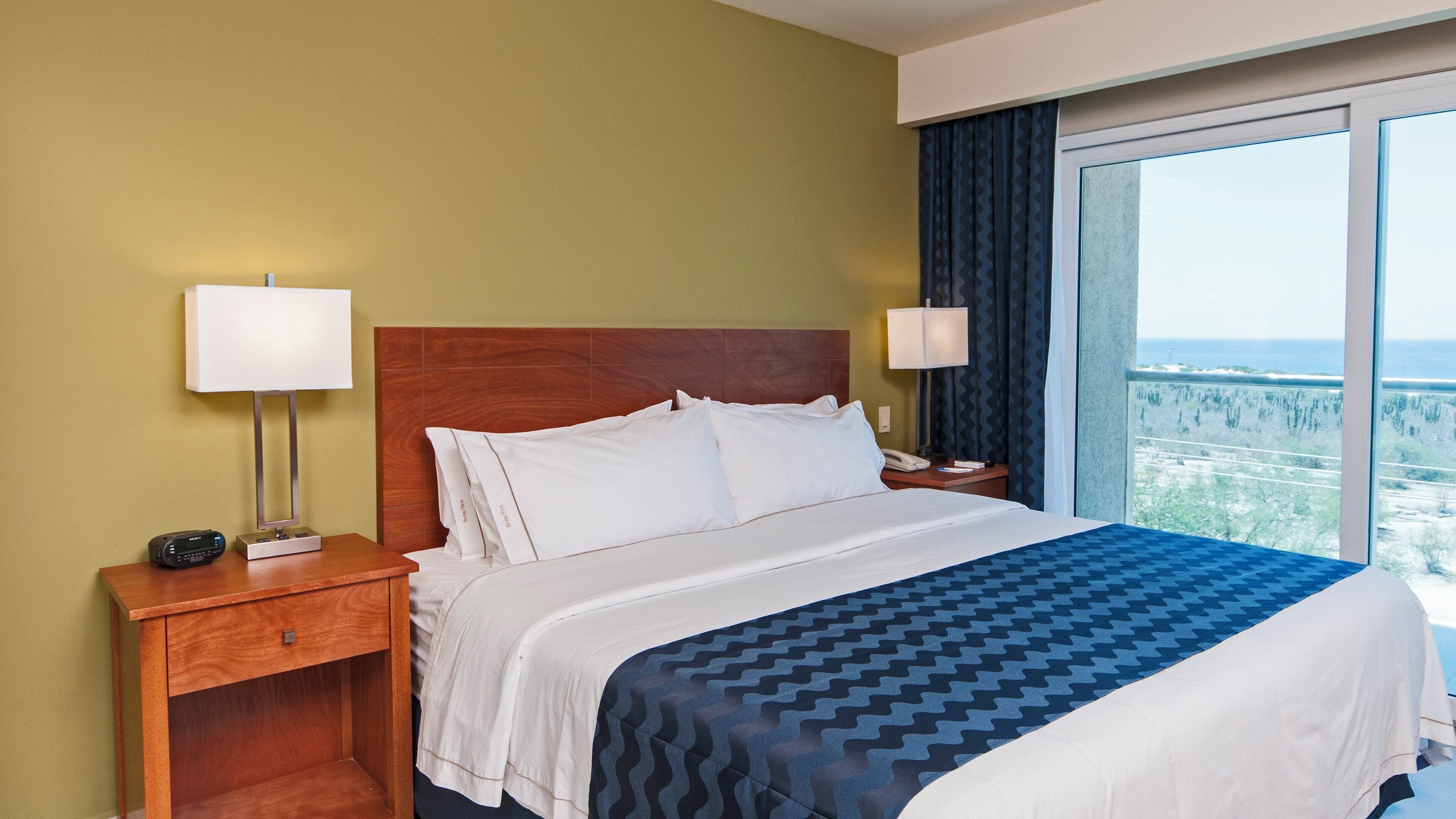 A hotel room with a bed, two bedside tables, a nightstand, a lamp, and a view of the outdoors.