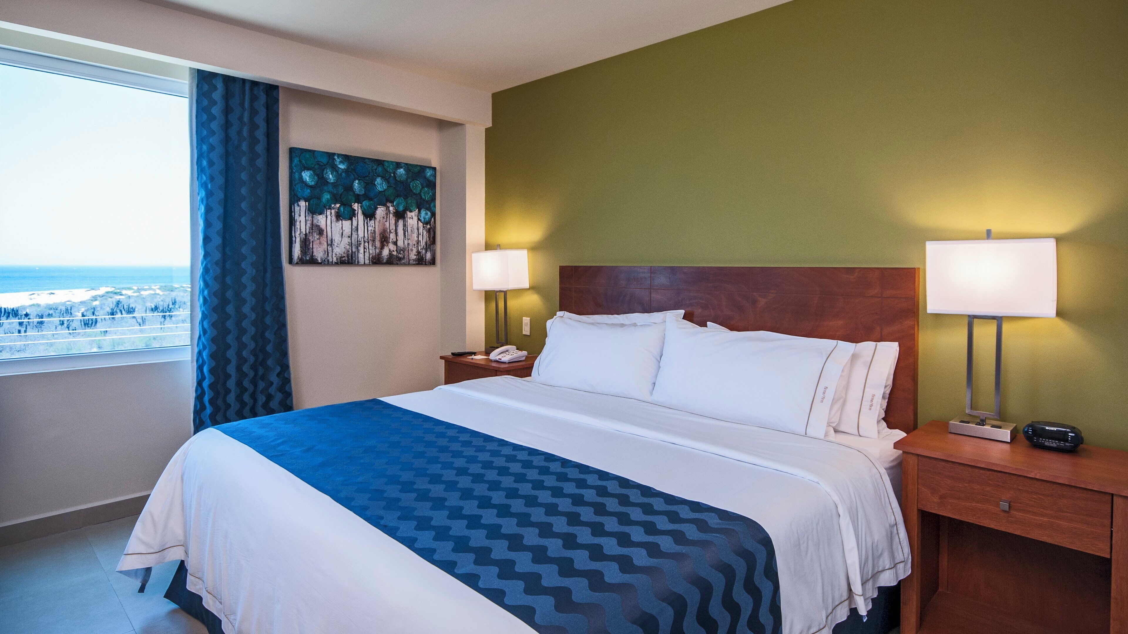 A hotel room with a bed, two bedside lamps, a nightstand, a painting, and a view of the ocean.