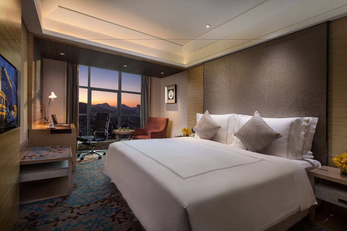 Une chambre d’hôtel avec un grand lit, un bureau avec une chaise, une petite table avec une lampe, et une vue sur les montagnes par la fenêtre.