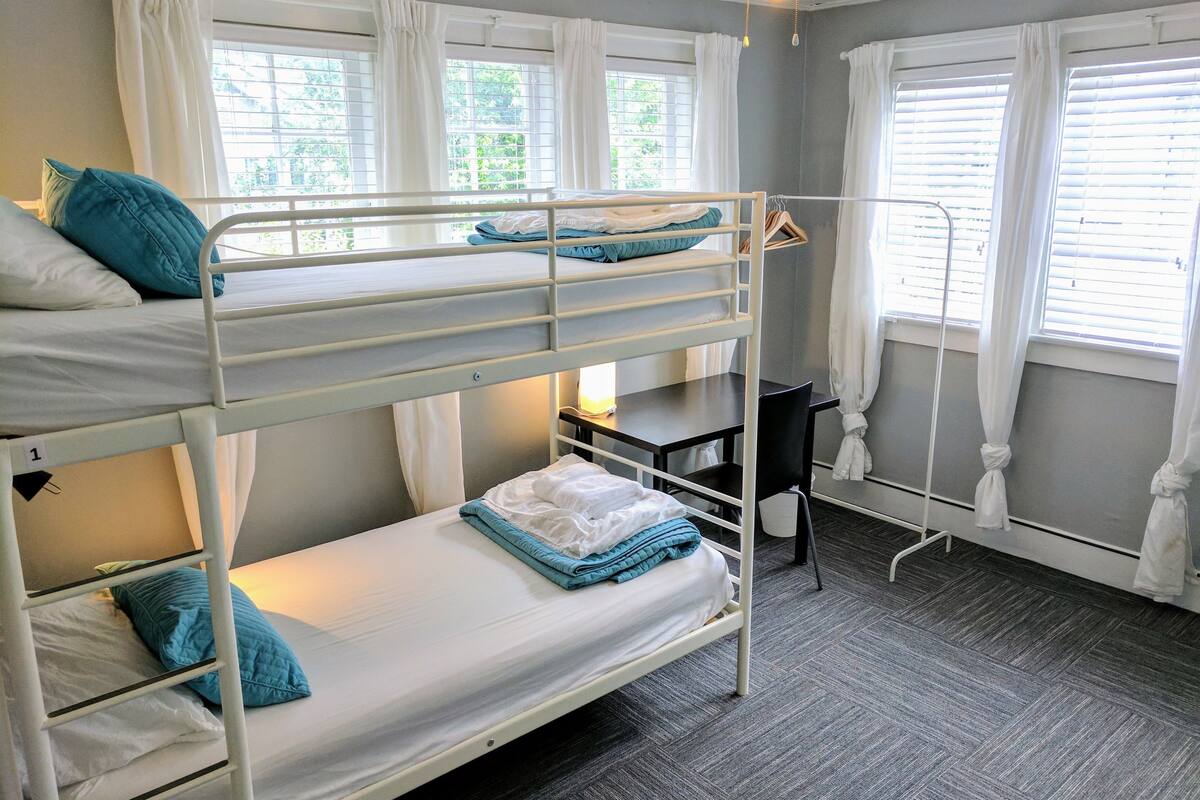 A bunk bed room with a desk and chair, white curtains, and a window with blinds.