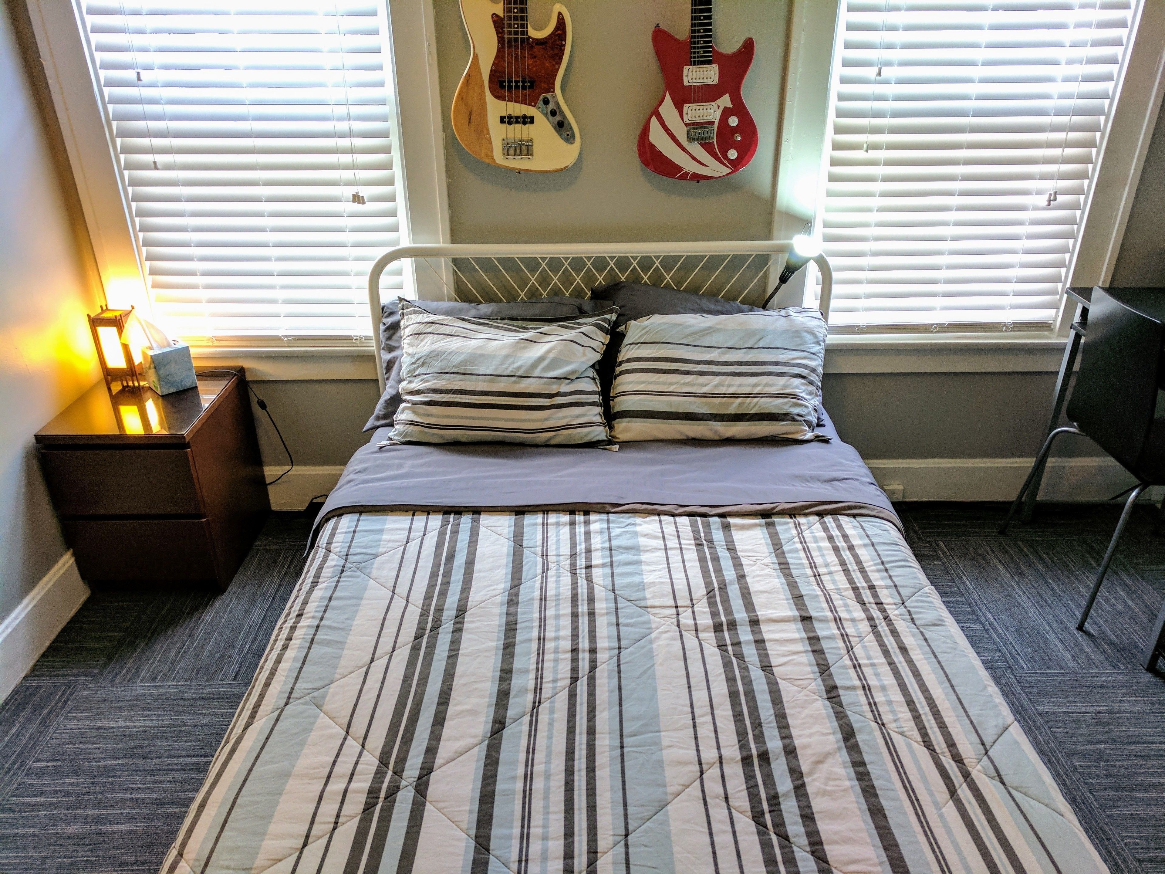 A bedroom with a bed, a nightstand, a lamp, two guitars on the wall, and a chair.
