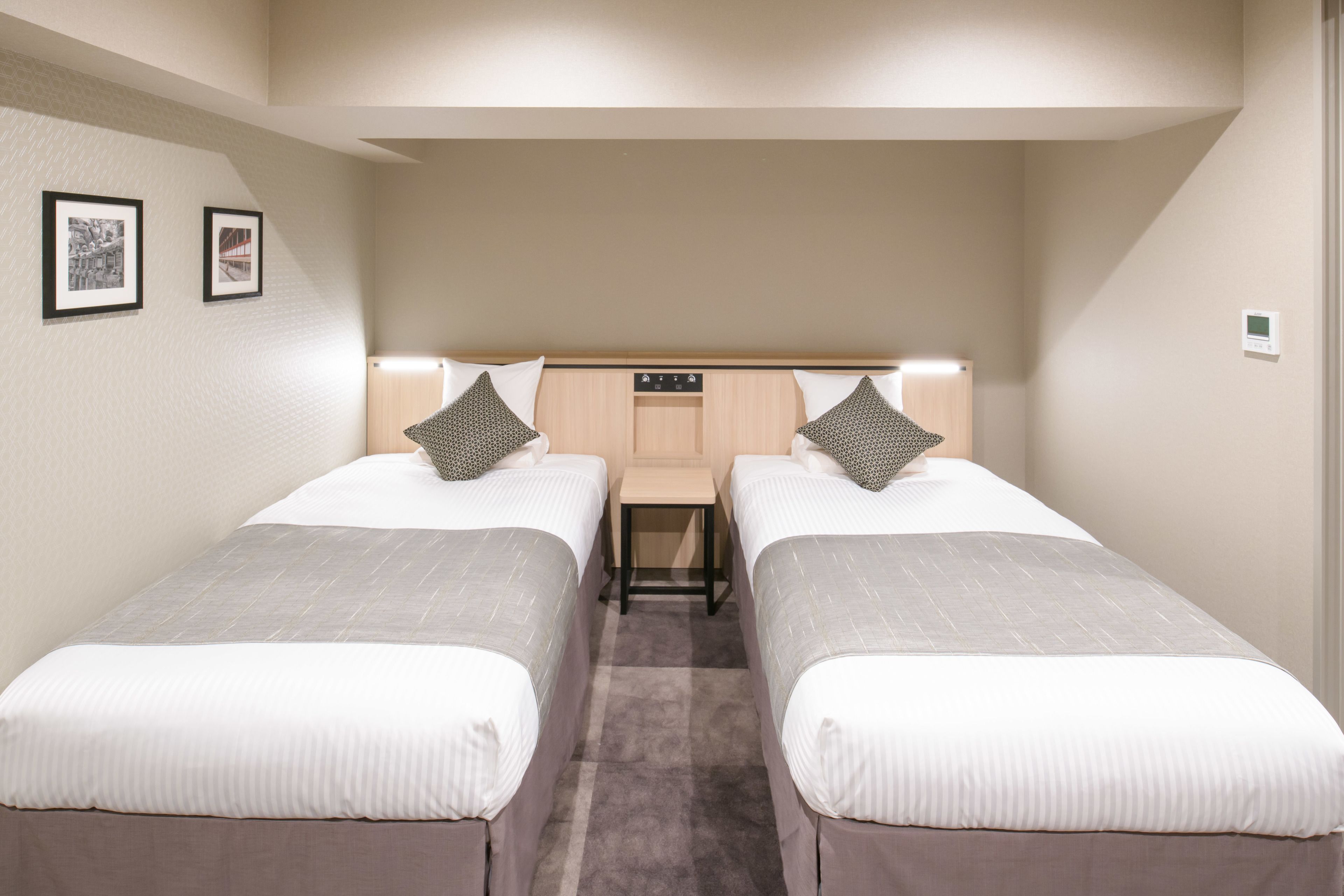 Habitación de hotel con dos camas individuales, una mesita pequeña y un cabecero con panel de control.
