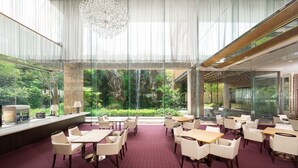 A spacious indoor dining area with tables and chairs, large windows offering views of greenery and a waterfall, and a modern chandelier. - Nara Royal Hotel (Nara)