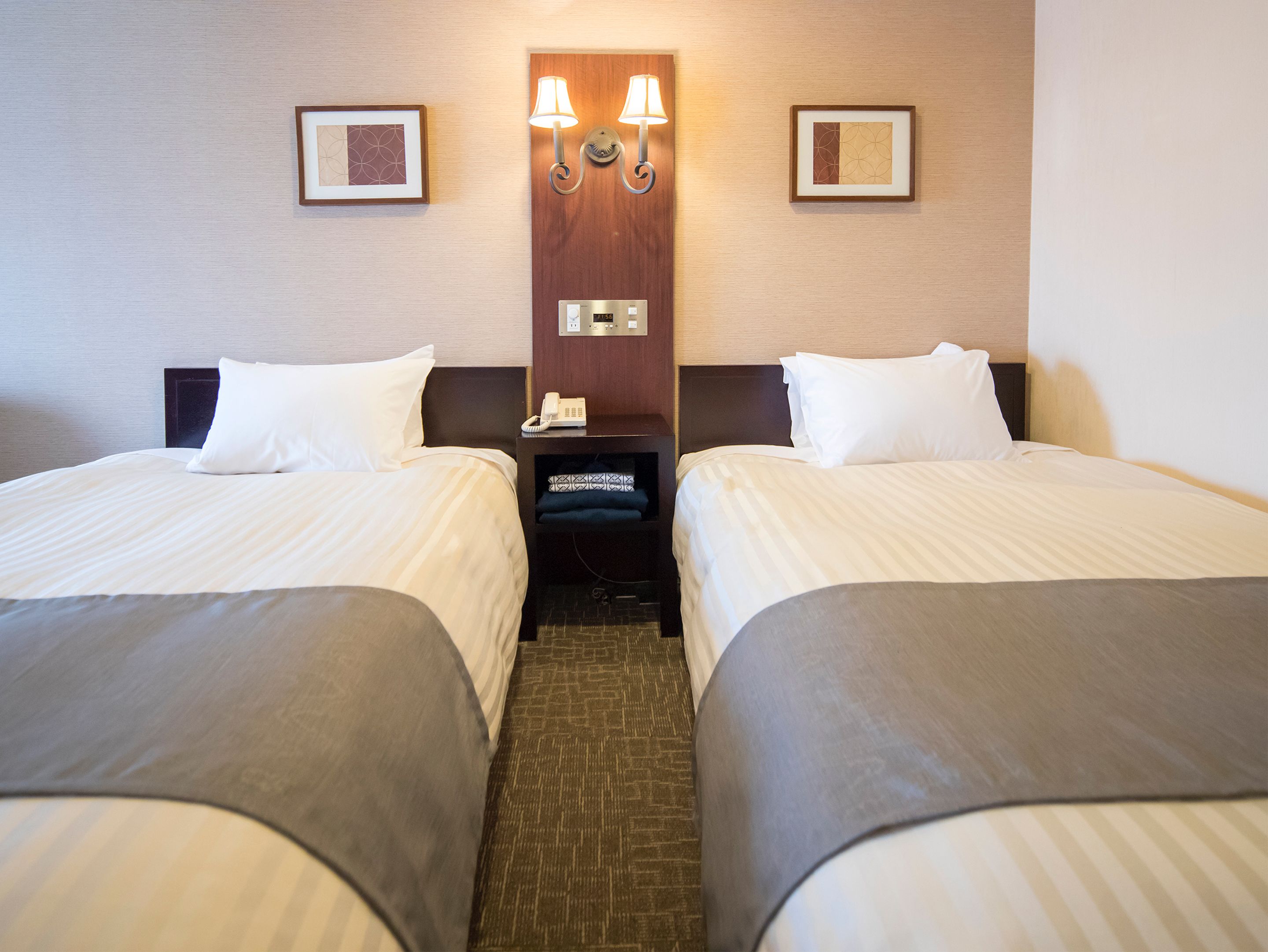 A hotel room with two beds, a nightstand with a phone, and wall-mounted lamps.