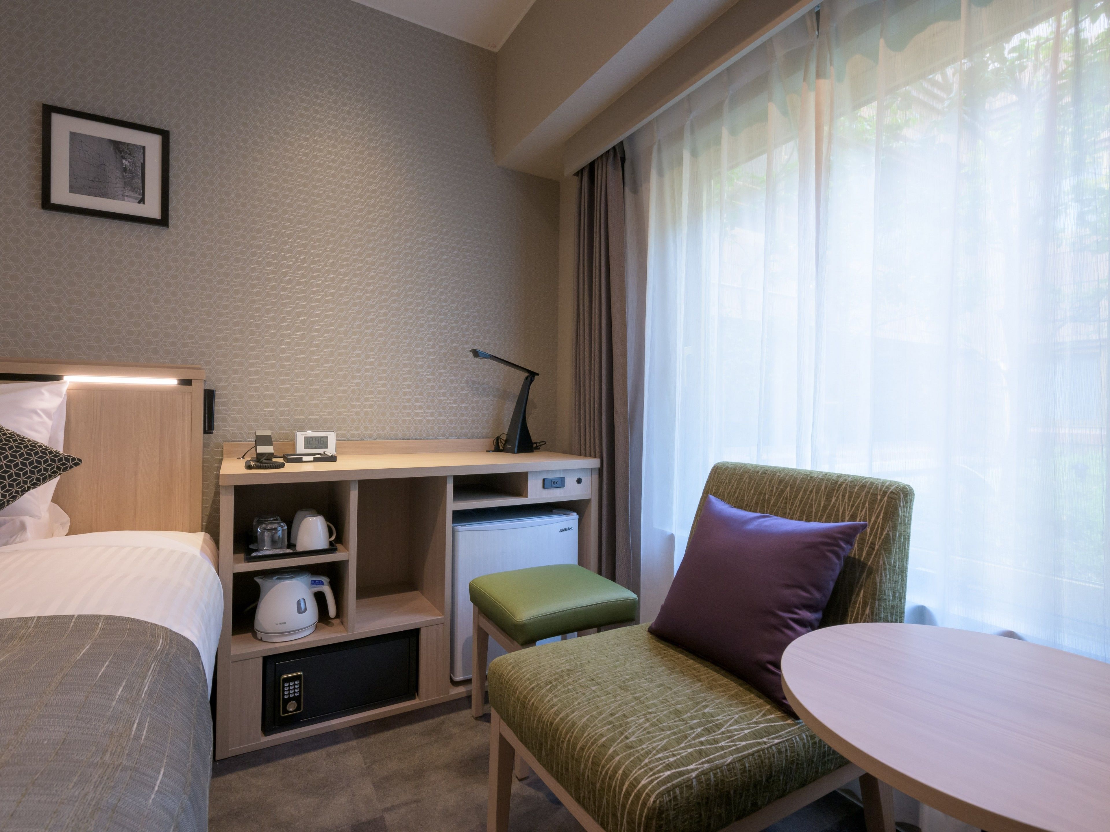 A hotel room with a bed, a desk with a computer, a chair, a small table, and a window with curtains.