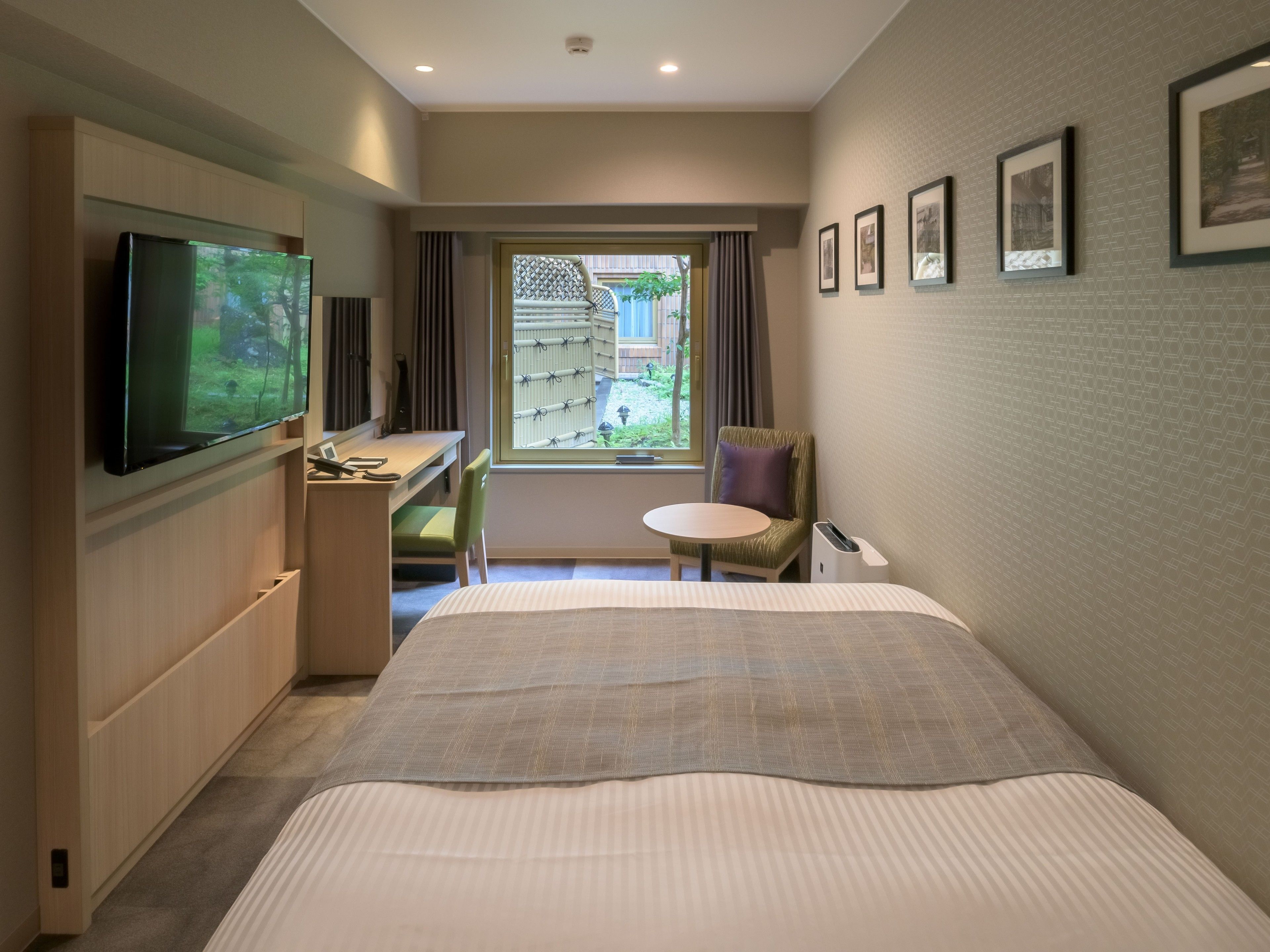 A hotel room with a large bed, a desk with a flat-screen TV, a chair, a small table, and a window with a view of the outside.