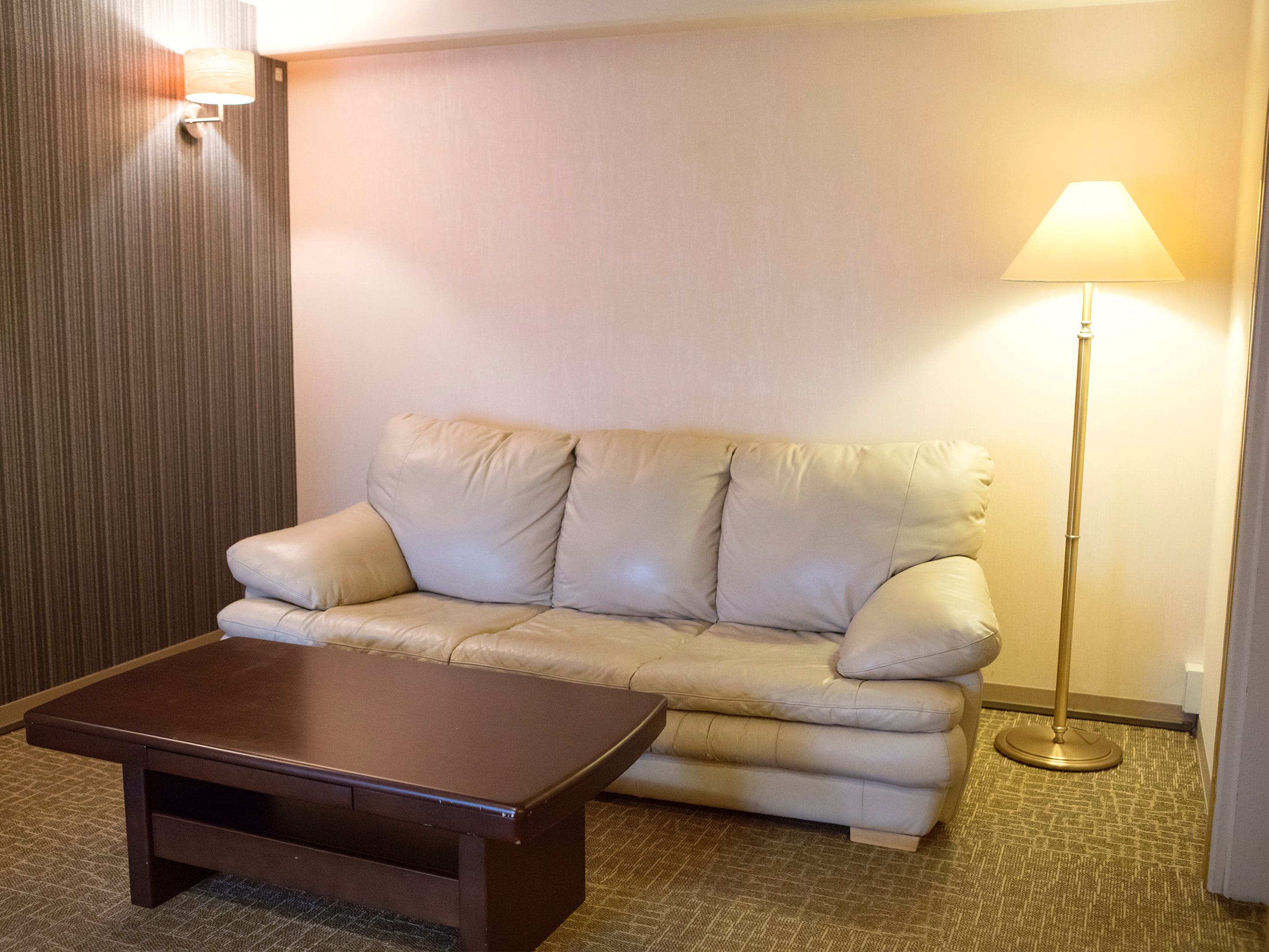 A beige leather sofa, a wooden coffee table, and a floor lamp in a hotel room.
