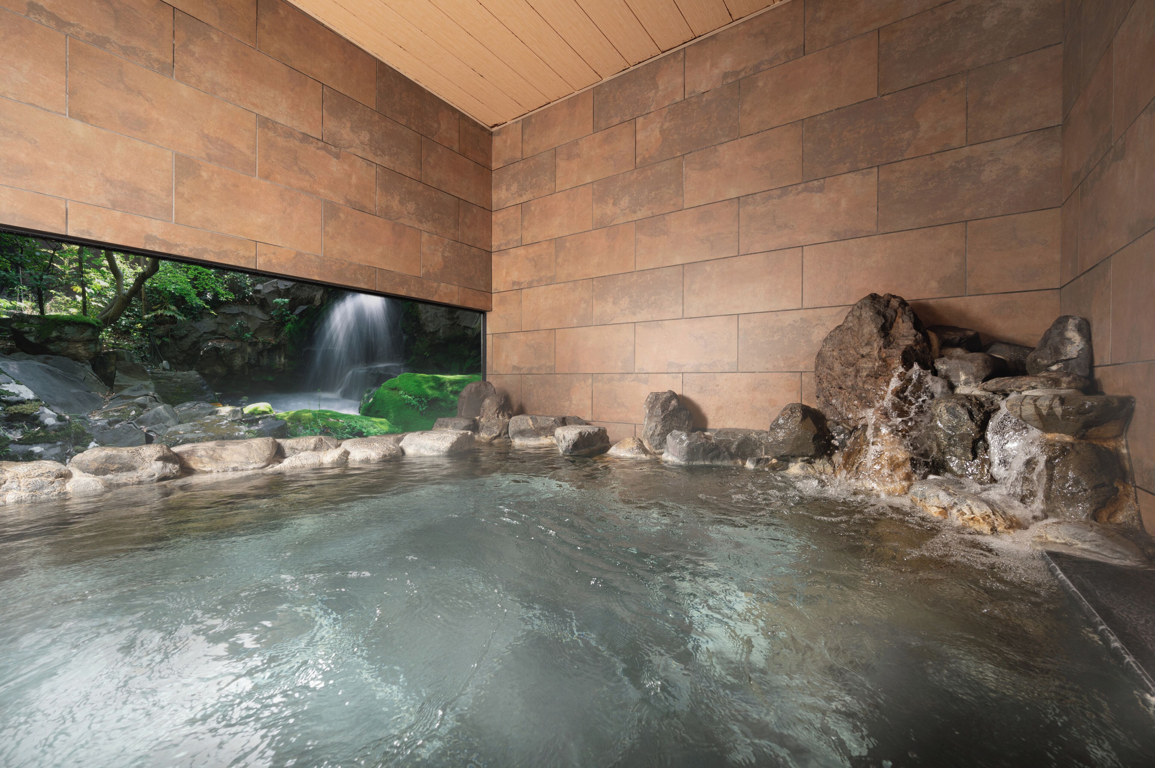 Ein Thermalbad mit Blick auf einen Wasserfall, Steinmauern und einer Holzbalkendecke.