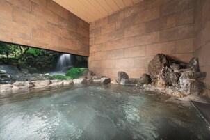 Un área de aguas termales con vista a una cascada, paredes de piedra y techo de madera.