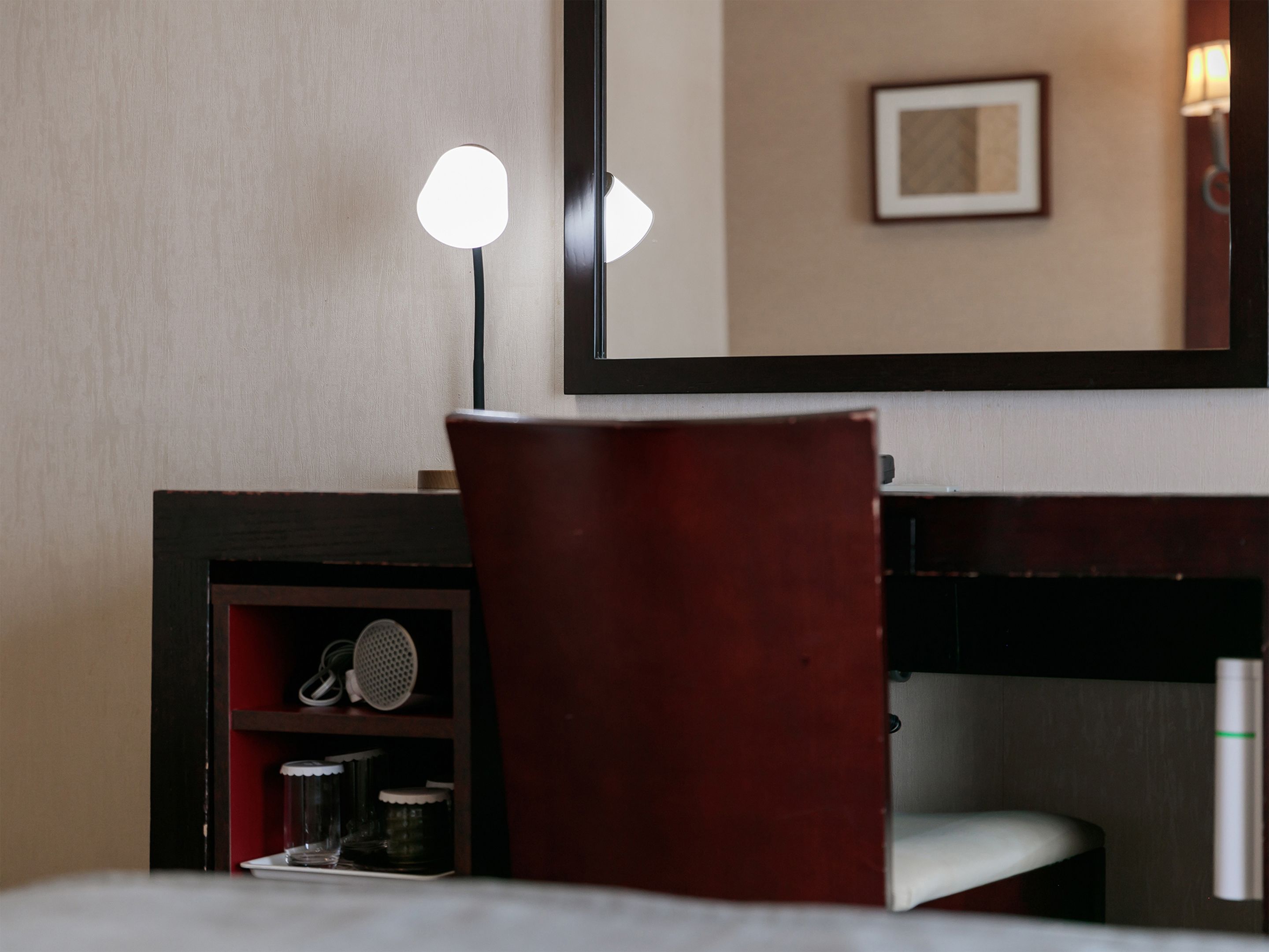 A hotel room with a bed, a desk with a chair, a mirror, and a shelf with a lamp and a small decorative item.