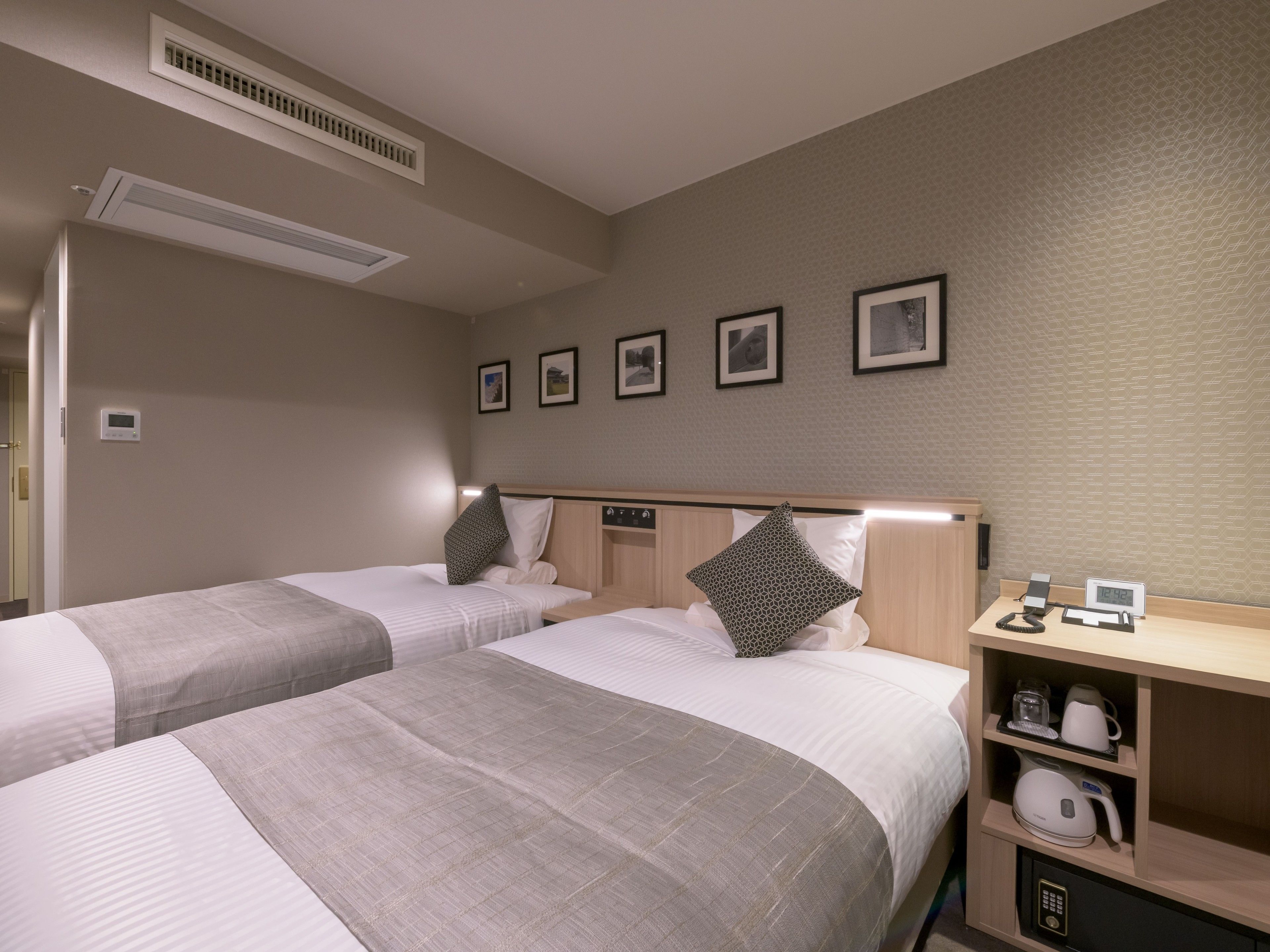 Ein Hotelzimmer mit zwei Betten, einem Nachttisch mit Kaffeemaschine, einer digitalen Uhr und einem Wandlüfter.